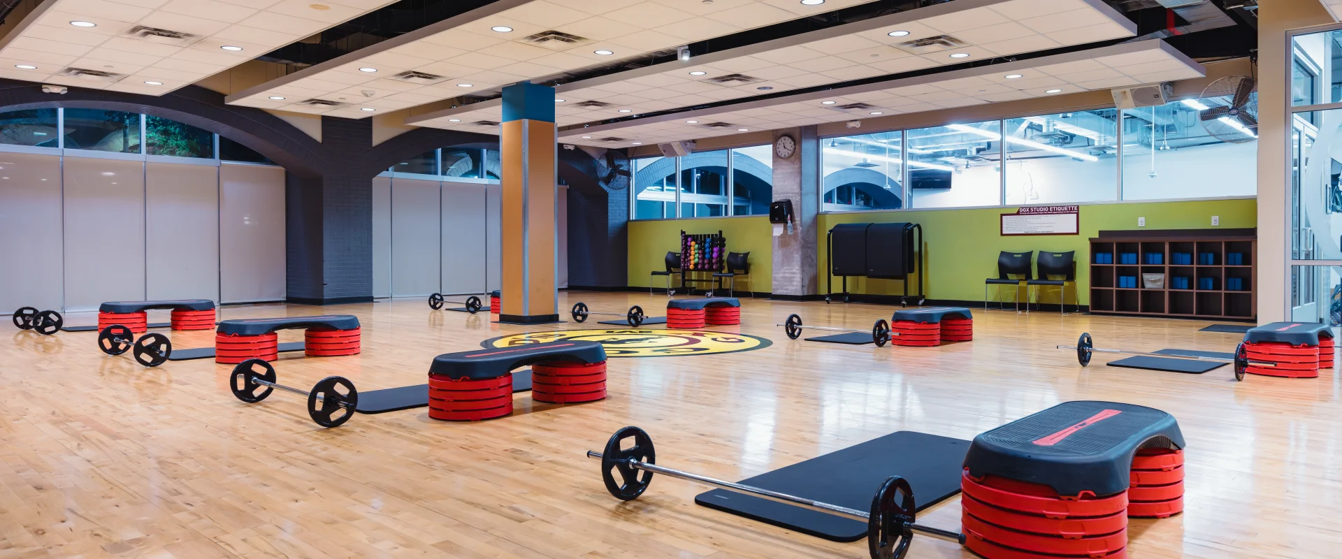 Group exercise studio at Gold's Gym Austin (Downtown) with polished wooden floors, floor-to-ceiling windows, and mirrored walls. The room is equipped with barbell sets, step platforms, and yoga mats arranged for a class.