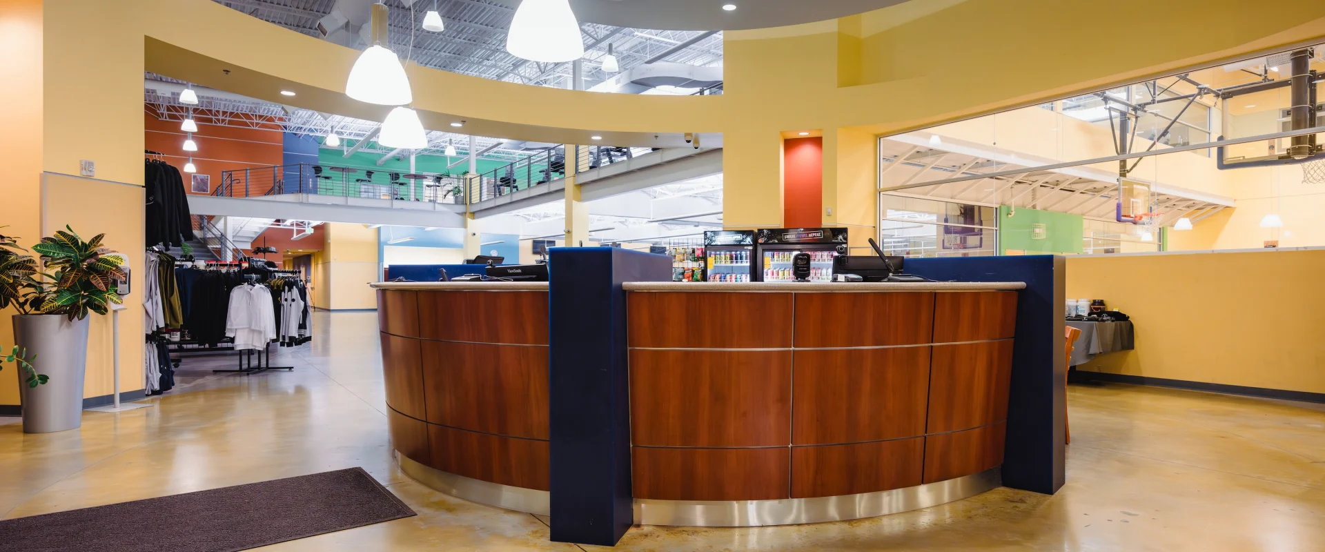 Modern gym reception area with a curved wooden desk, pendant lighting, adjacent basketball court, and merchandise display on the left. Bright and spacious ambiance.