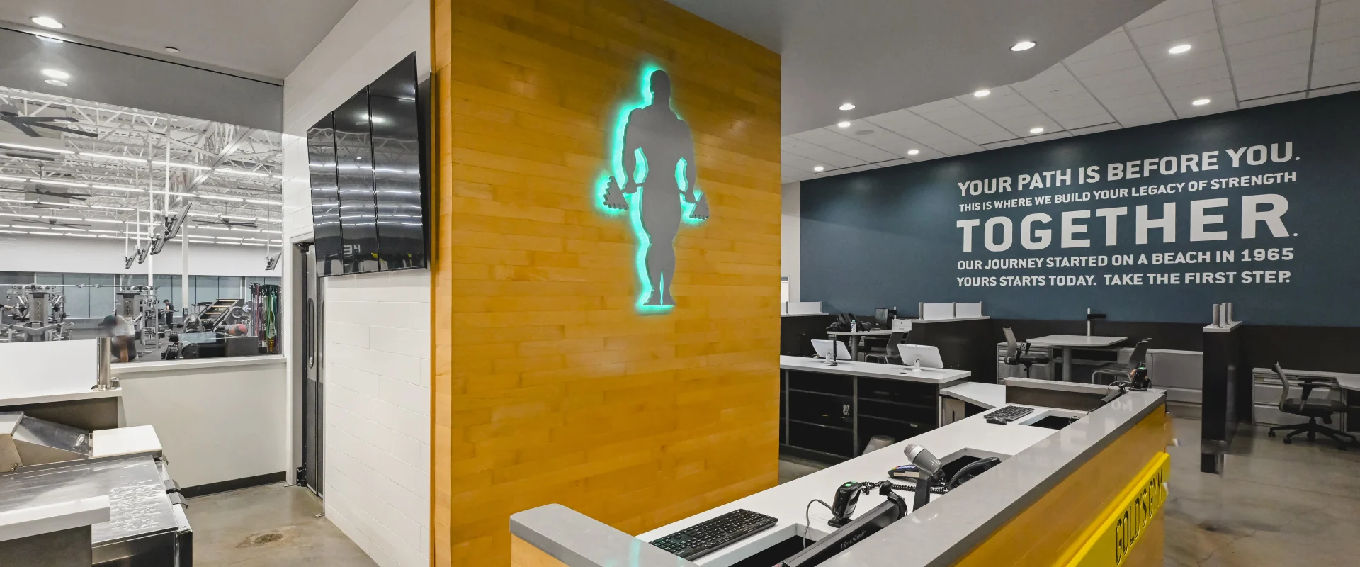 Front desk area at Gold's Gym College Station (Tower Point) featuring a sleek reception counter with a gray stone top and wood paneling. A glowing silhouette of a weightlifter decorates the wooden accent wall, while a motivational quote is displayed on a dark teal wall in the background.