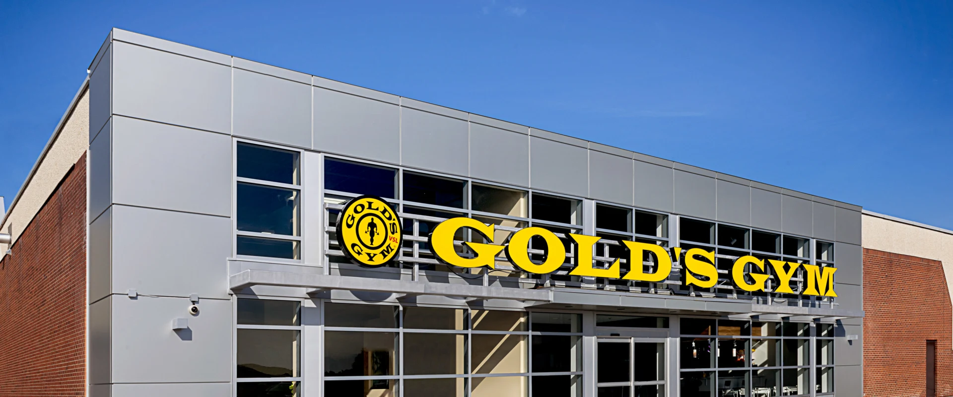 Exterior view of Gold's Gym Vertu with modern silver and brick facade, bold yellow Gold’s Gym signage, and large front-facing windows under clear blue skies.