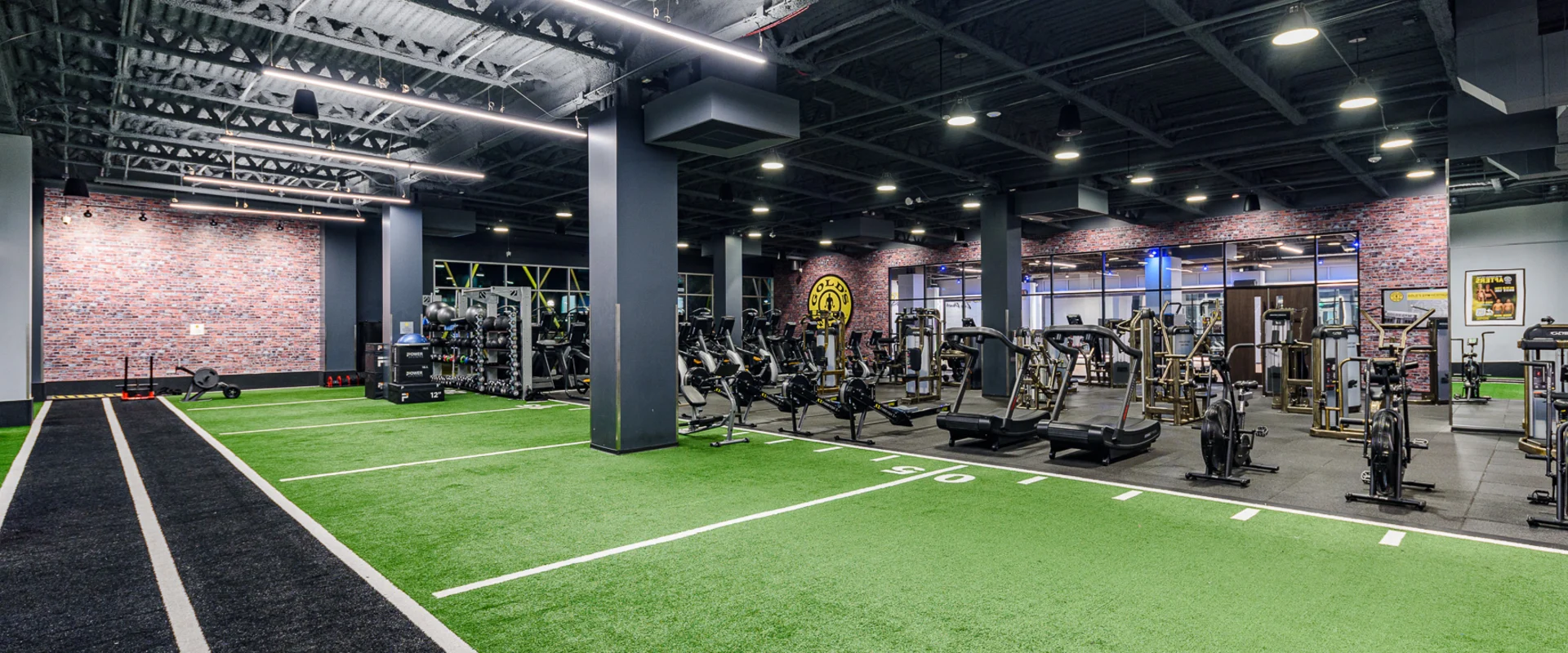 Turf area at Gold’s Gym Long Beach, designed for functional training and athletic performance. The space includes green turf lanes for sled pushes, agility drills, and sprint work, alongside a black track for conditioning exercises. Racks stocked with kettlebells, battle ropes, and medicine balls line the perimeter. Adjacent to the turf, a variety of cardio machines, rowers, and strength equipment provide additional training options. The open layout, industrial lighting, and exposed ceiling beams contribute to the gym’s modern, high-energy atmosphere.