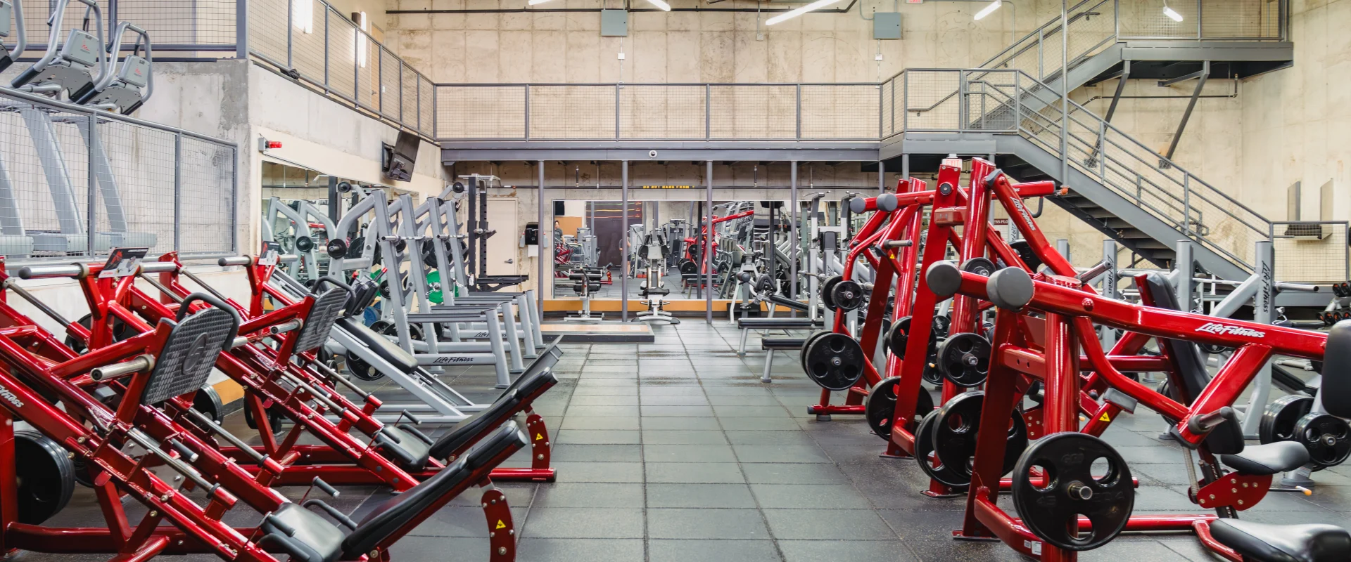 Strength training area at Gold's Gym Austin (Downtown) featuring plate-loaded machines and benches with bold red frames. The industrial-style space includes concrete walls, modern overhead lighting, and rubber flooring designed for durability and comfort.