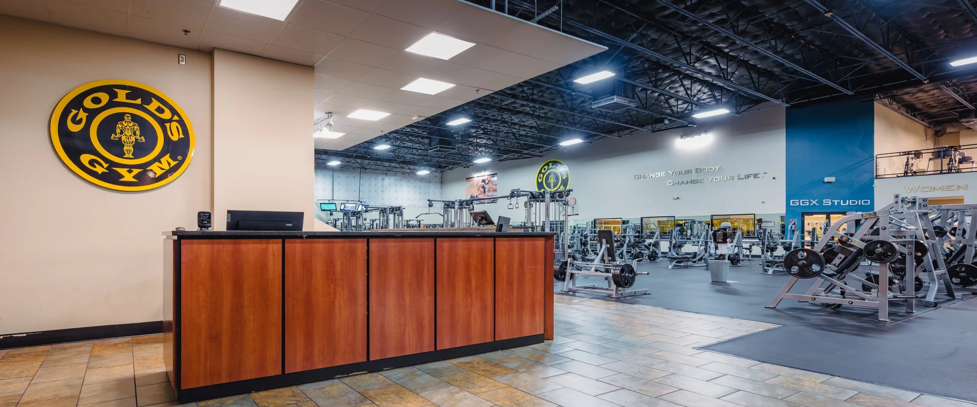 The front desk at Gold’s Gym in Austin (South) features a sleek wooden counter with a black granite top, set against a beige wall adorned with the bold Gold’s Gym logo. The tiled floor in earthy tones adds warmth to the entryway. Beyond the desk, the spacious gym floor is visible, filled with strength training equipment and free weights. Bright overhead lighting highlights the open layout, while a blue accent wall marks the GGX Studio entrance. Motivational quotes on the walls, such as “Change Your Body. Change Your Life,” add inspiration to the space.