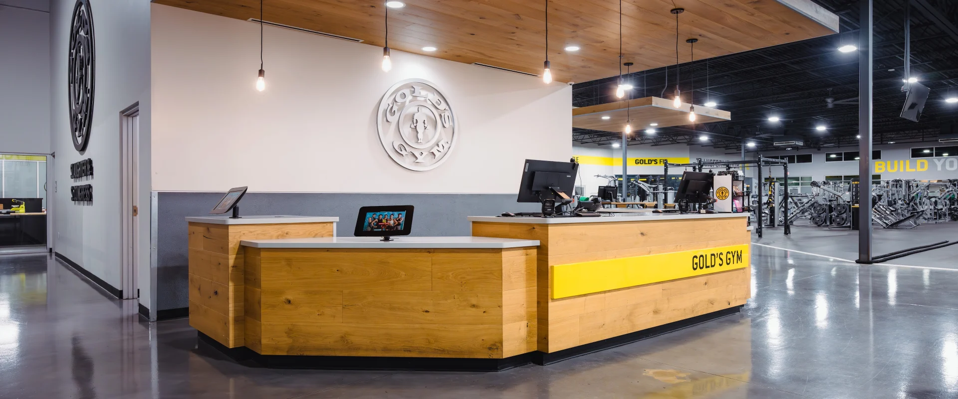 The front desk at Gold's Gym in Texas (Slaughter & South Congress) features a wood-accented counter with modern lighting fixtures overhead. Check-in stations are equipped with digital tablets, while the gym’s logo is prominently displayed on the wall behind the desk.
