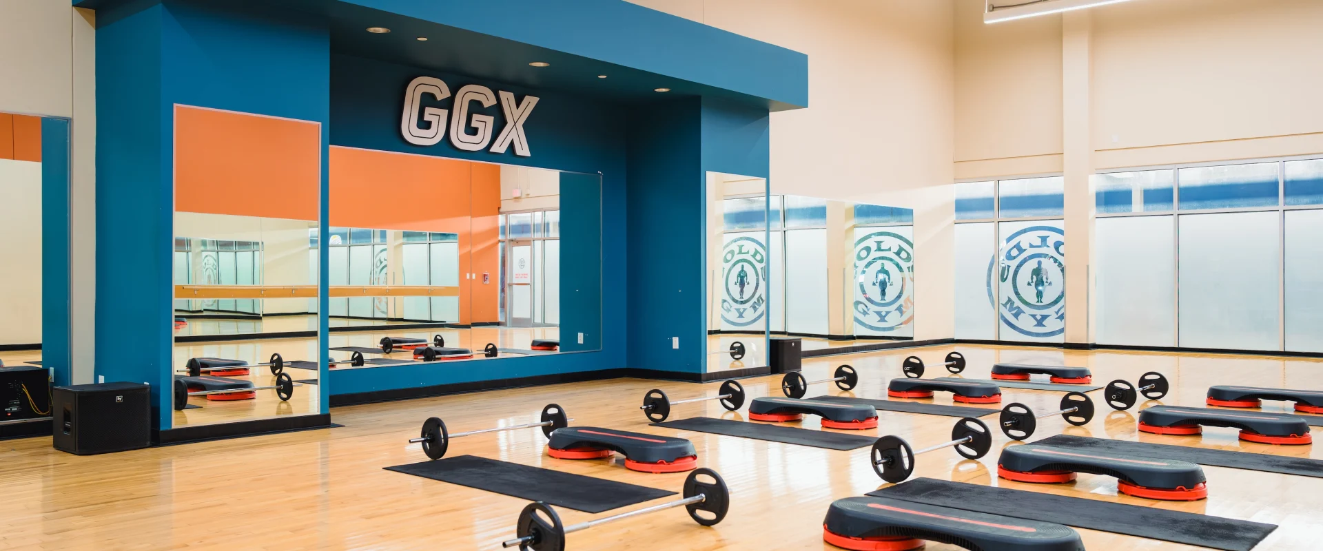 Spacious fitness studio with wooden floors, black mats, weights, and red platforms. Mirrors line the wall under a blue structure labeled “GGX.” Bright, organized setting.
