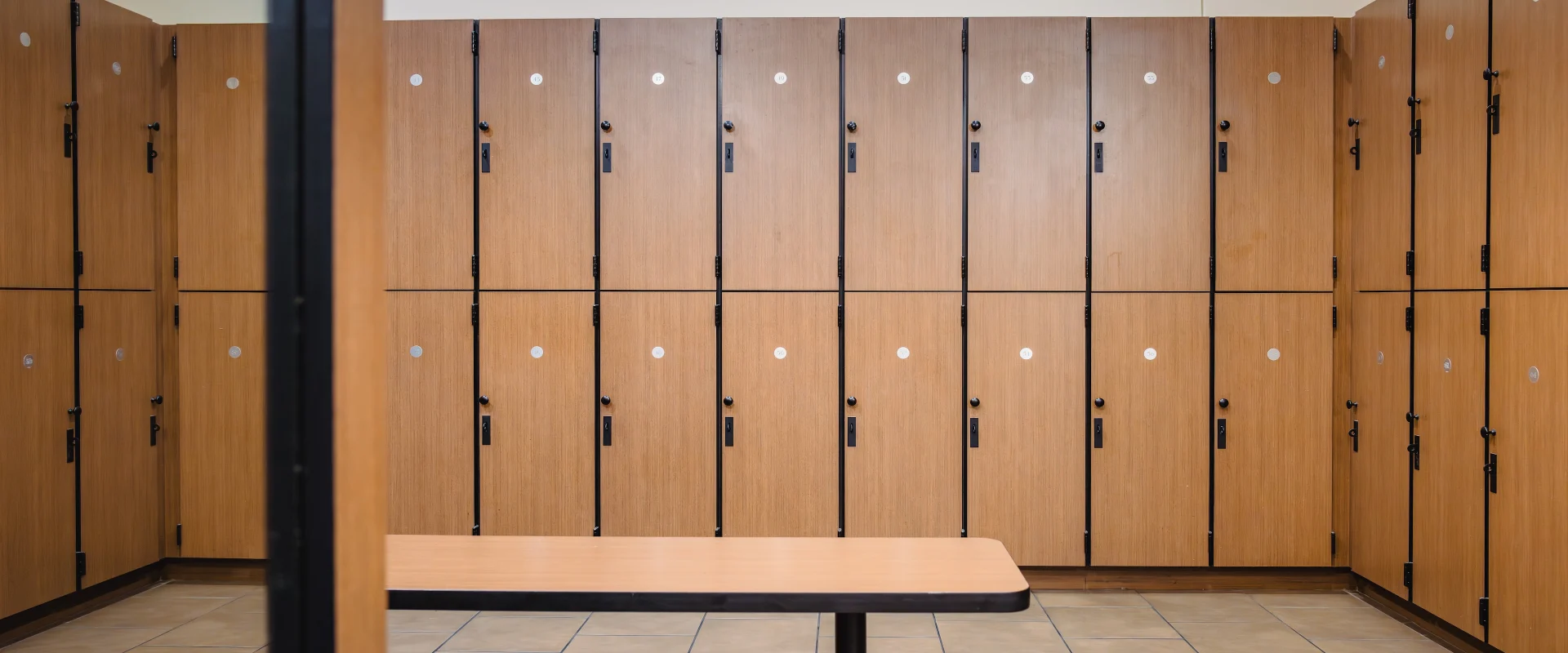 Locker room in Austin (Westlake), featuring rows of wooden lockers with black trim and secure locking mechanisms. The space includes tiled flooring and a central wooden bench with a sturdy black metal base. The clean, minimalist design promotes a functional and comfortable environment.