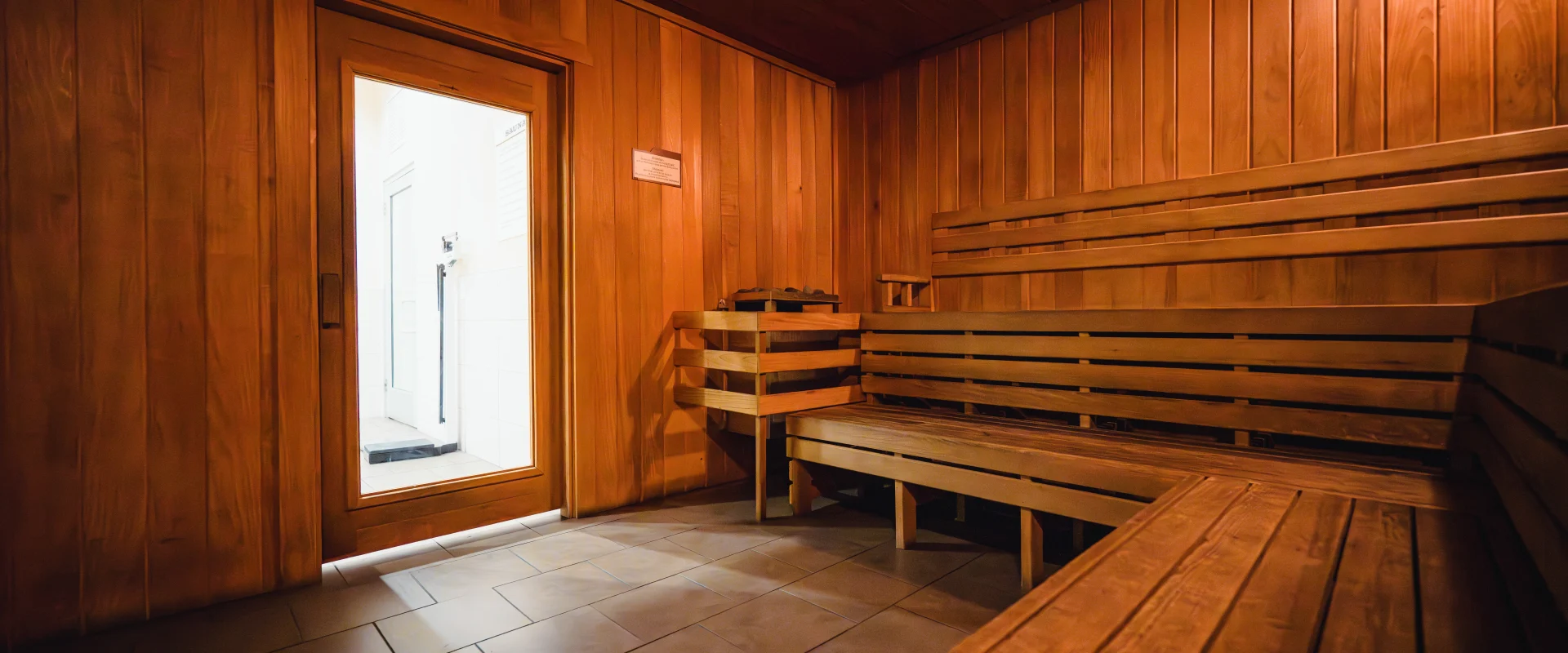 Interior of a wooden sauna at Gold's Gym, featuring warm wood paneling, bench seating, and a door with a window leading to a brightly lit exterior area.