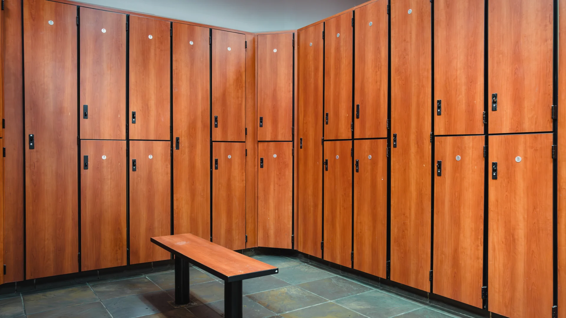 Locker room at Gold's Gym Waco featuring wood-paneled lockers with black accents, a matching wood bench, and a tiled floor with a natural stone look.