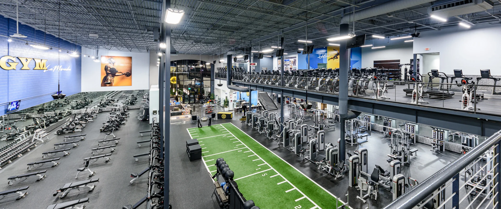 An expansive view of the gym floor from an elevated perspective. Rows of free weights, resistance machines, and a turf area dominate the space. The Gold’s Gym logo is displayed on the blue walls, with cardio machines lining the upper deck.