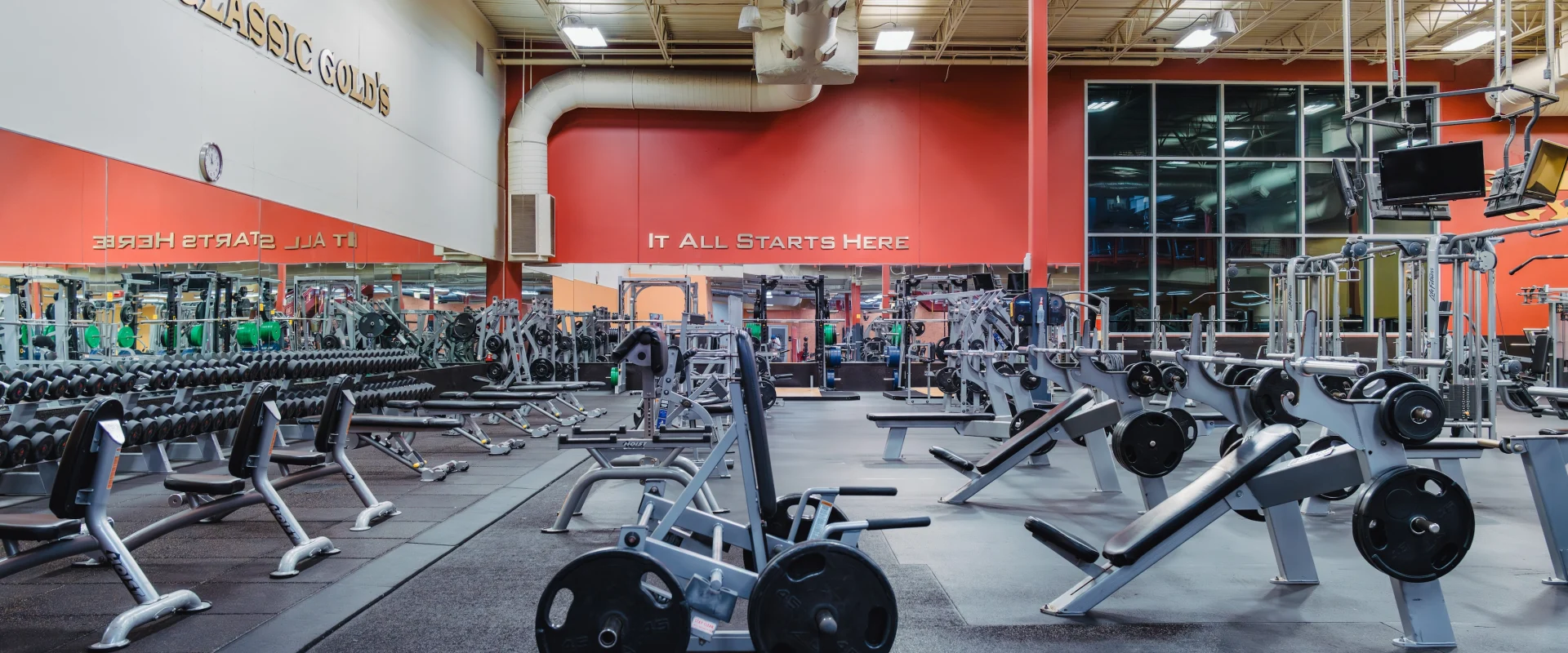 Gold's Gym Fiesta Trails interior with red and white walls, weightlifting equipment, large windows, and mirrors.
