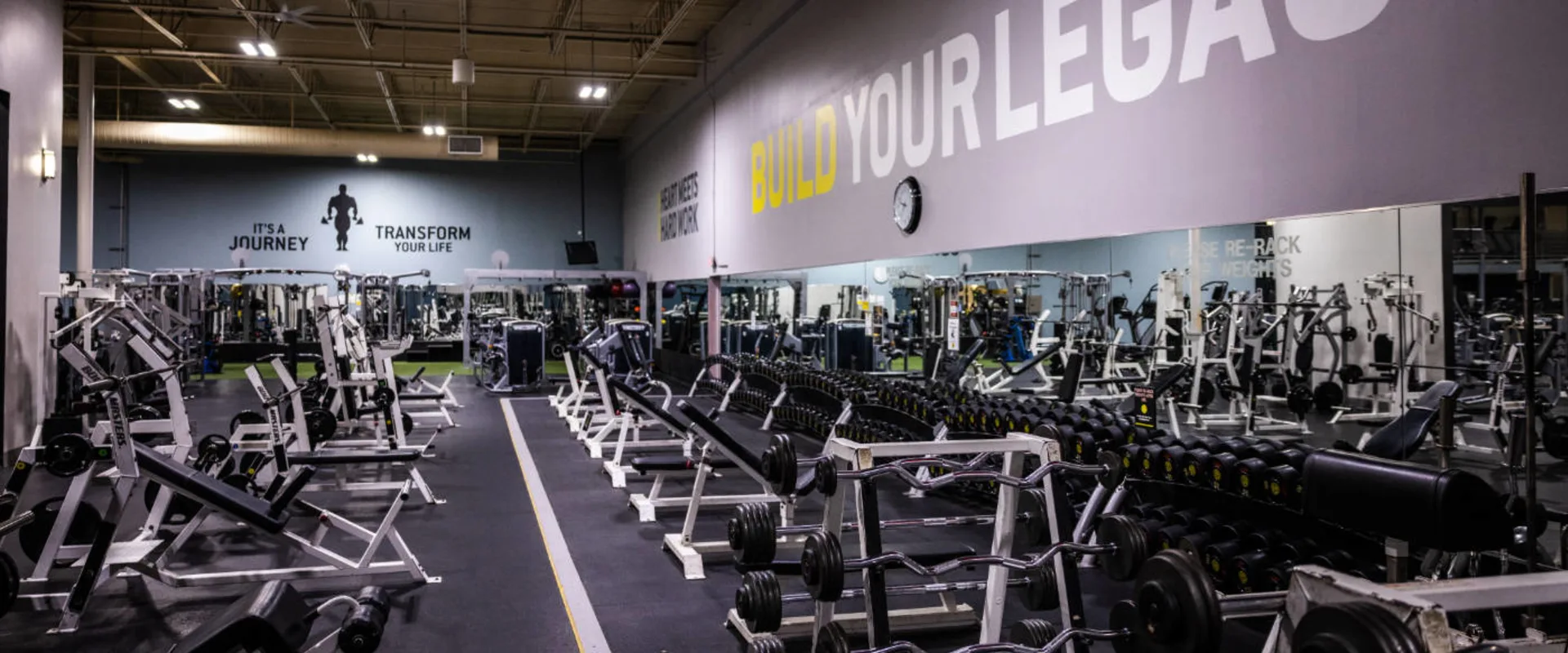 A free weights area at Gold’s Gym in Woodbridge (Lake Ridge) features multiple rows of dumbbells neatly arranged on racks against a mirrored wall. Barbells and EZ curl bars are placed on a separate rack in the foreground. Several white and black weightlifting benches are positioned throughout the space. A yellow and white "BUILD YOUR LEGACY" sign stretches across the mirrored wall, with a clock mounted above. In the background, additional strength training machines and squat racks extend into the gym, with large motivational graphics on the walls. Bright overhead lighting illuminates the space.