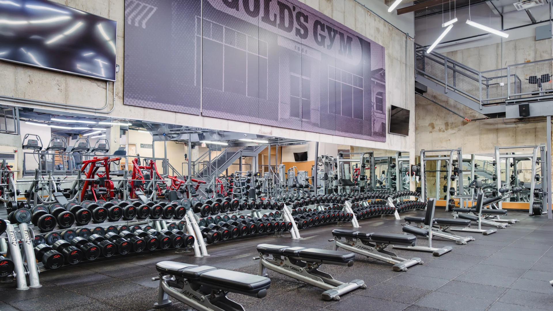 Interior of Gold's Gym Austin (Downtown) with rows of dumbbells on racks, adjustable weight benches on rubber flooring, mirrored walls reflecting gym equipment, industrial-style light fixtures, a wall-mounted TV, and an old photo of Gold's Gym Venice displayed above the mirrors.