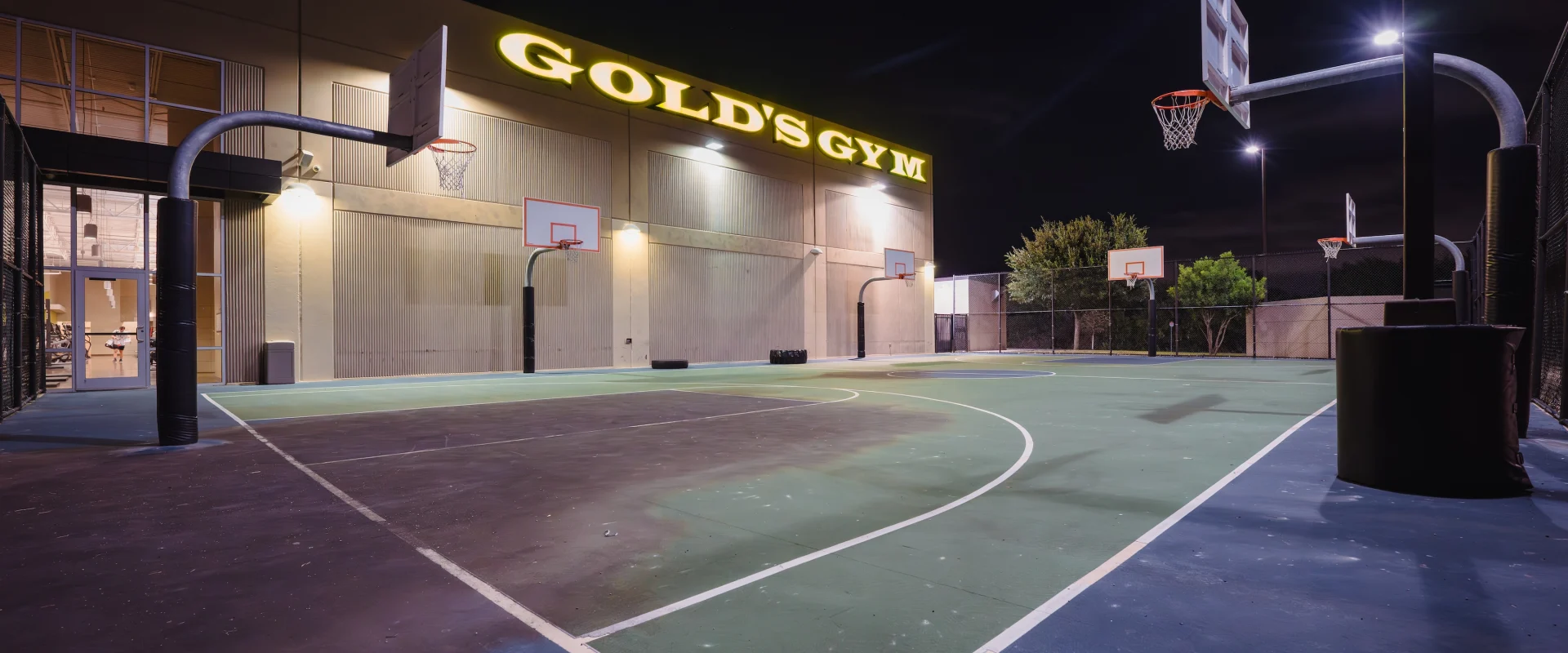 Outdoor basketball court at night, lit by overhead lights, with "Gold's Gym" sign on a building. The scene is calm and inviting.