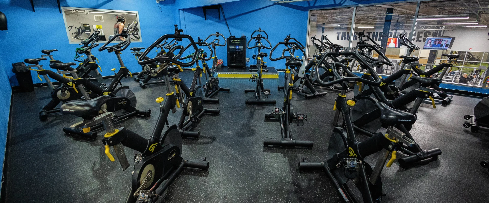 Group cycle studio at Gold’s Gym in Fishkill, New York, featuring rows of stationary bikes with yellow accents arranged for spin classes. The space is enclosed with vibrant blue walls and large windows overlooking the main fitness floor. A mounted TV and audio equipment enhance the class experience, while the dark rubber flooring provides stability and comfort for high-intensity cycling sessions.