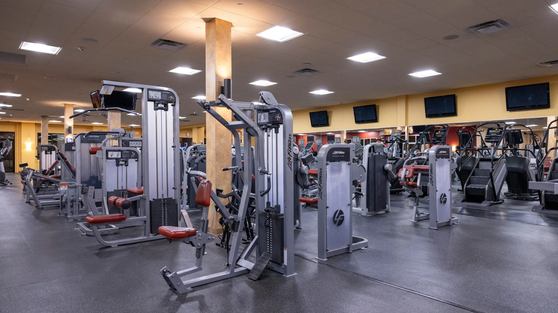 Strength equipment area at Gold’s Gym in East Northport, New York, featuring Life Fitness selectorized machines for targeted muscle training, including leg extension, bicep curl, and chest press machines. The space is organized with rubber flooring, mirrored walls, and overhead TVs lining the perimeter. Bright ceiling lights enhance visibility, while Matrix stair climbers are visible in the background for cardio integration.