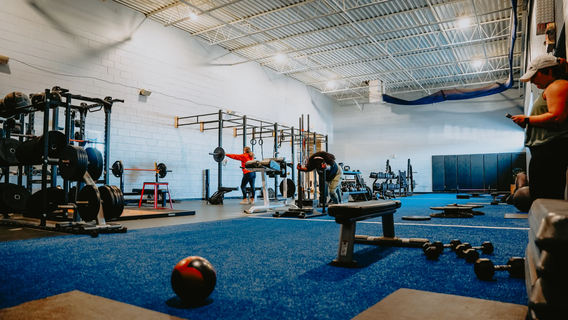 Functional training area at Gold’s Gym Tennessee, Lee Highway, featuring a wide-open space with vibrant blue turf flooring. The area includes racks of weights, barbells, and functional equipment like gymnastic rings and medicine balls. A variety of functional stations and benches are arranged across the space, with members engaged in exercises. The white walls and high ceilings with exposed beams and bright overhead lighting create an airy, focused workout environment.