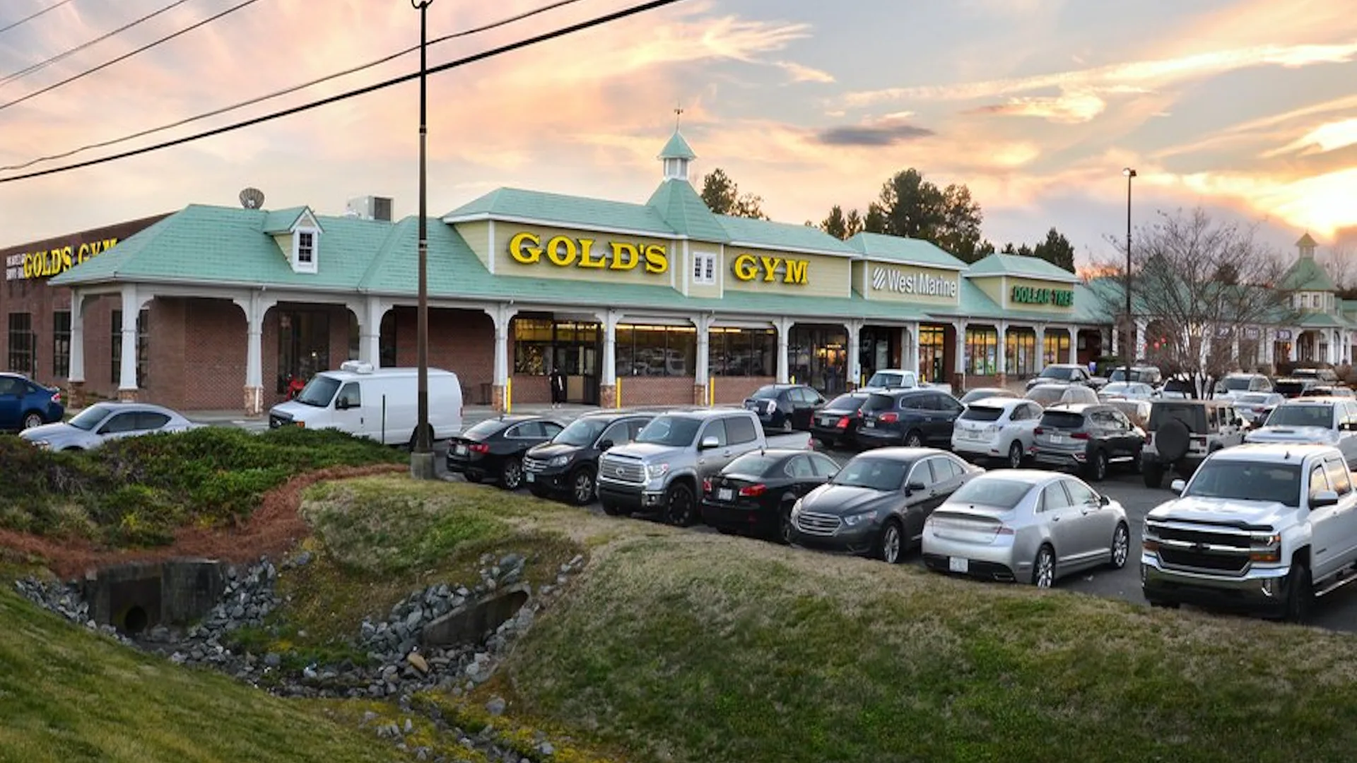 Exterior of Gold’s Gym Lake Norman with yellow signage and green roof in shopping plaza at sunset.