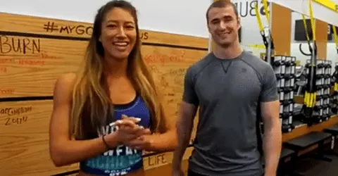 The image shows two people, a woman and a man, standing together in what appears to be a gym or fitness center setting, with various exercise equipment and signage visible in the background.