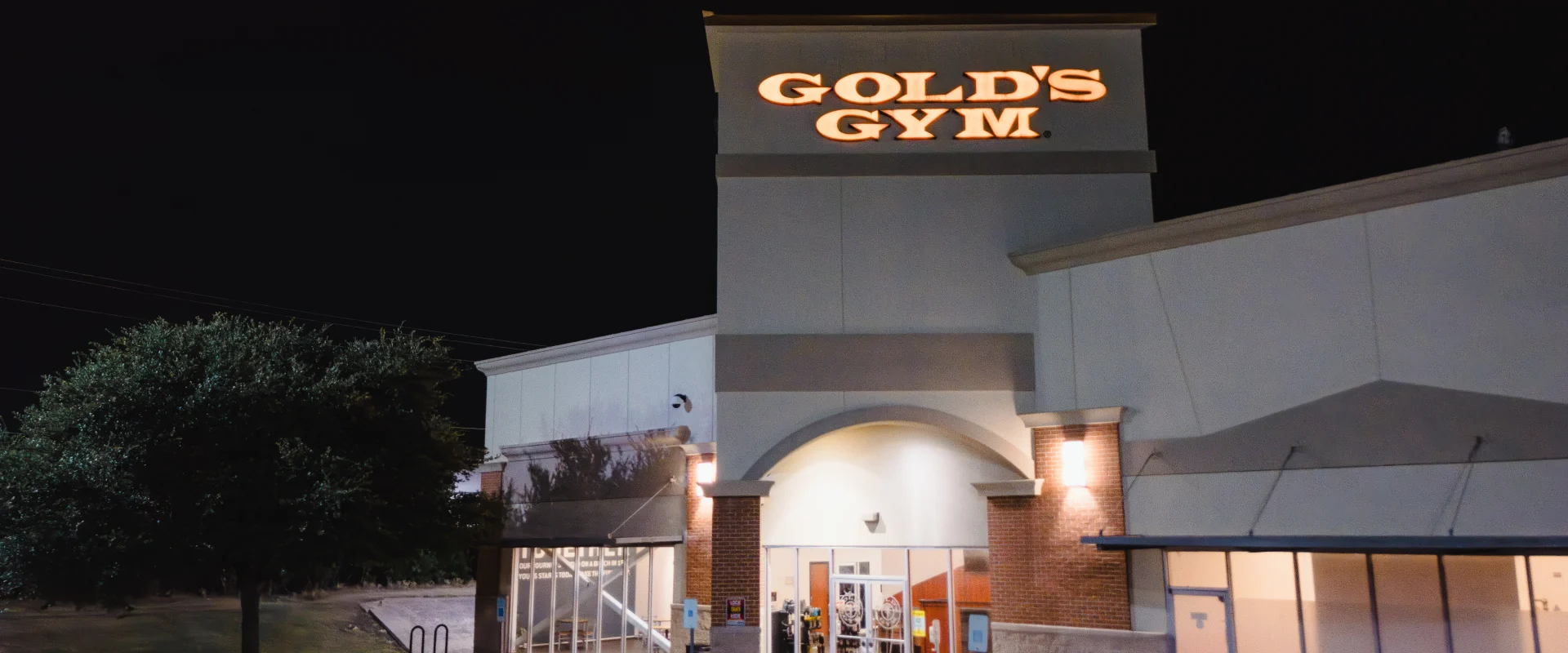Gold's Gym exterior at Austin (Hesters Crossing) captured at night, featuring a well-lit building facade with an illuminated logo. The entrance is framed by arched architecture and large glass windows, while the parking area in the foreground is dimly lit, with a tree visible to the left adding contrast to the scene.