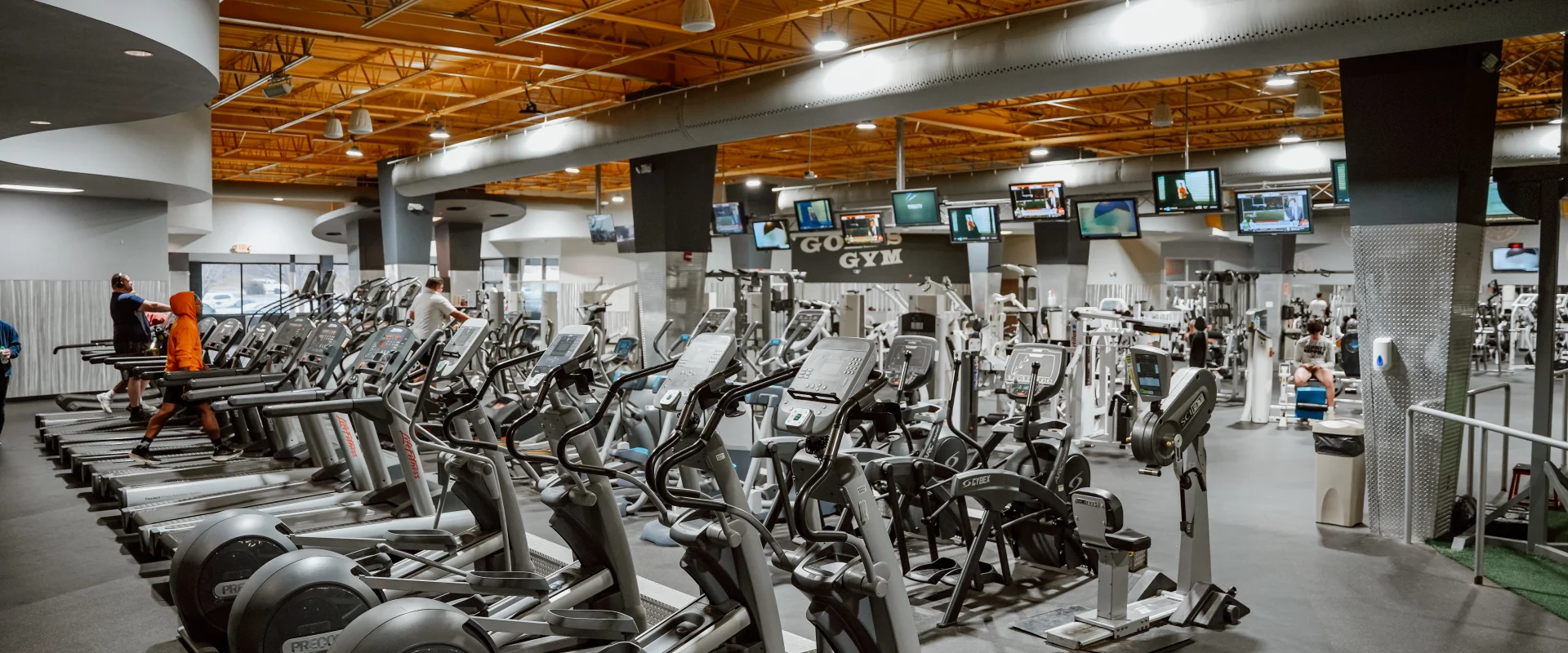 Cardio equipment area at Gold’s Gym, Chapman, Tennessee, featuring rows of treadmills and elliptical machines in use by members. Overhead, flat-screen TVs display various programs, adding a dynamic and engaging atmosphere. The space is well-lit with industrial-style fixtures and a bright yellow ceiling, which contrasts with the modern gray walls and equipment. Additional gym equipment is visible in the background, highlighting the spacious and fully equipped facility.
