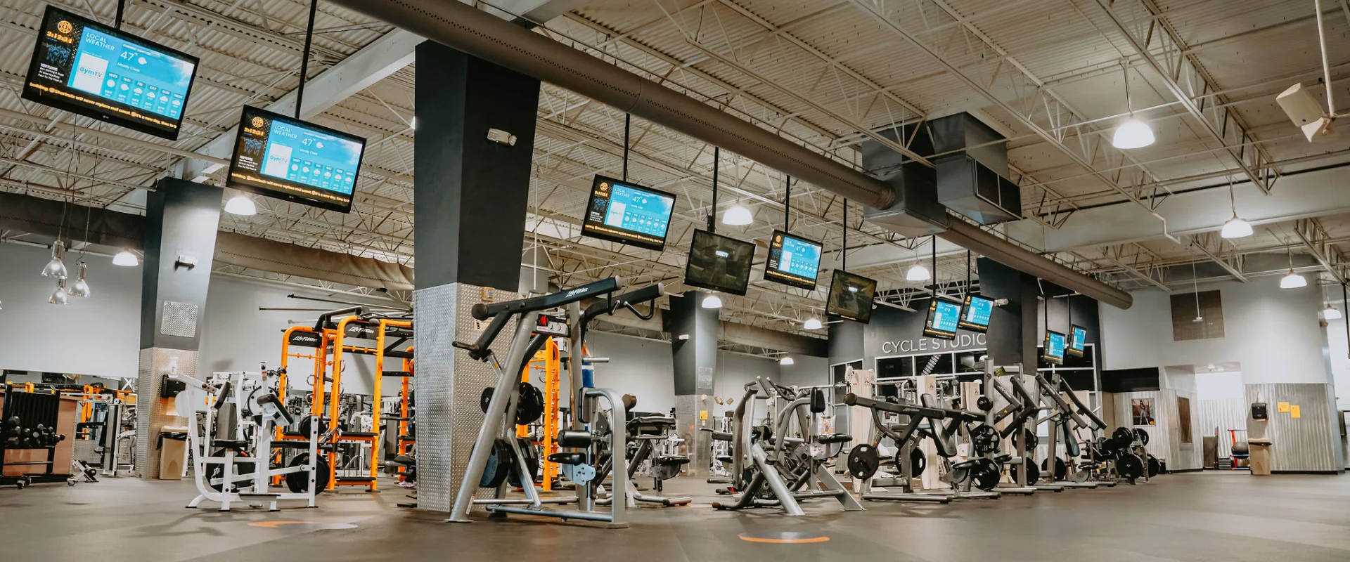Machine weight area at Gold’s Gym Tennessee, Hixson, featuring various strength training machines arranged on a dark rubber floor. The space includes multiple overhead screens, high ceilings with exposed ducts, and industrial-style lighting. A Cycle Studio entrance is visible in the background, alongside additional weight racks and gym equipment.