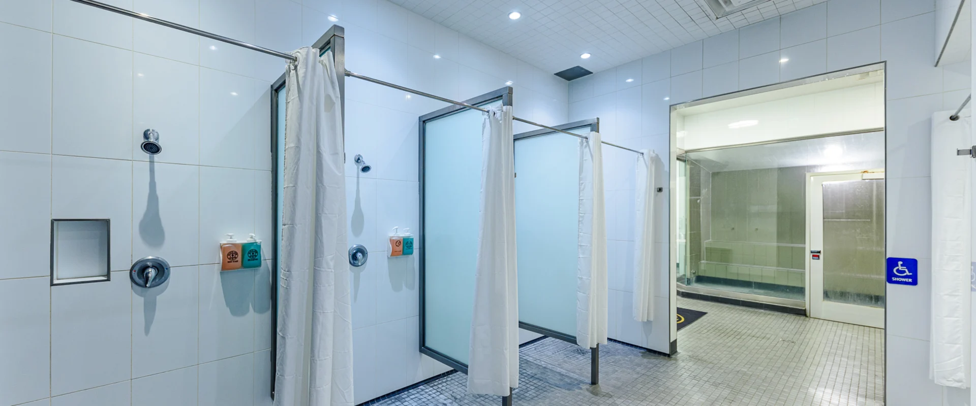Shower area at Gold’s Gym Hollywood, featuring multiple individual shower stalls with frosted glass dividers and white curtains. The walls are covered in large white tiles, while the floor consists of small square mosaic tiles with central drainage. Each shower includes a wall-mounted showerhead, a built-in shelf, and dispensers for soap and shampoo. An accessible shower stall is marked with a blue sign. In the background, a steam room with a glass door and tiled interior is visible. Recessed ceiling lights provide bright illumination.
