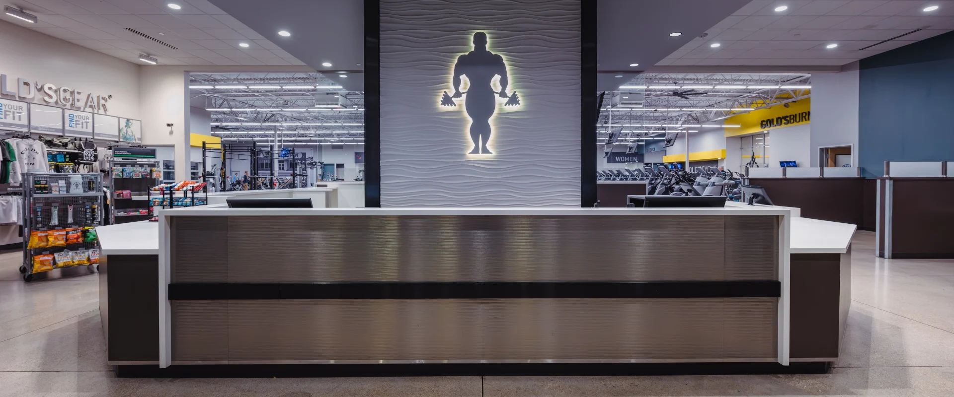 Front desk at Gold's Gym in Texas (Pflugerville), featuring a sleek, modern reception counter with a metallic finish. A backlit silhouette of a bodybuilder is centered on a textured wall behind the desk. The surrounding area showcases gym equipment, the Gold's Gear pro shop, and clear views of the workout floor under bright, spacious lighting.