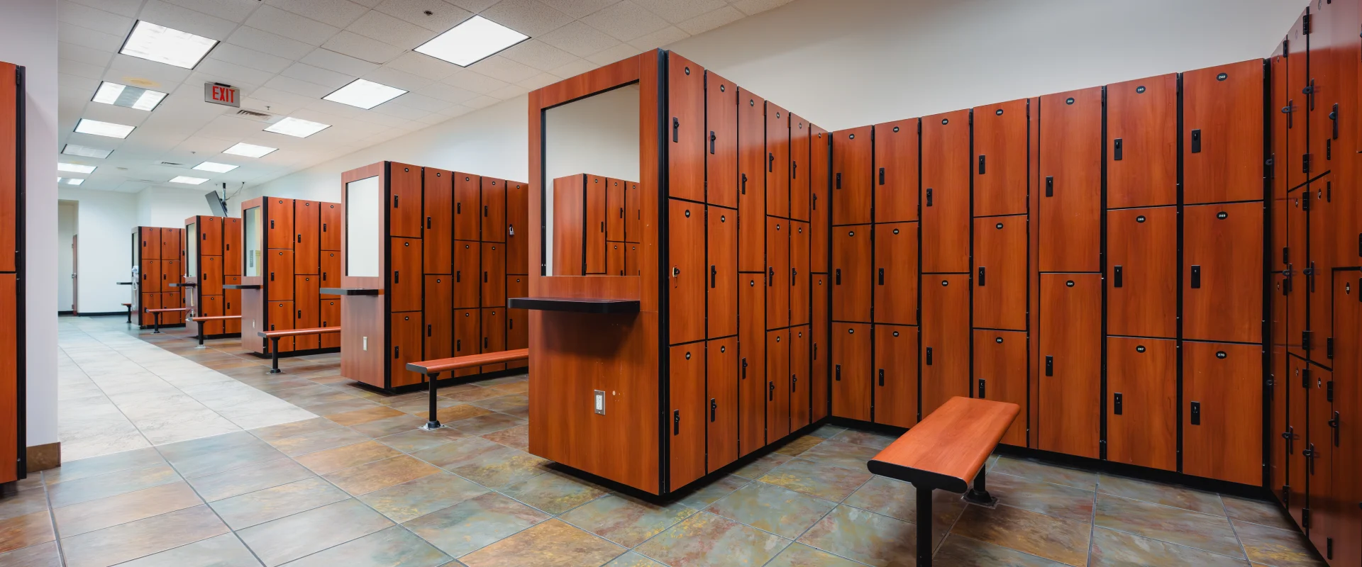 Locker room with rows of warm-toned wood lockers featuring black accents. Benches with matching wood tops and metal legs are positioned between the locker units. The room has a tiled floor in earthy tones, a drop ceiling with bright recessed lighting, and a clean, spacious layout. A wall-mounted fan is visible near the ceiling.