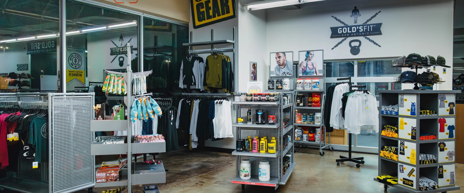 A well-organized Pro Shop with fitness gear and apparel. Racks display clothing, shelves hold supplements and gear, and decorations include motivational posters and signs. The atmosphere is vibrant and energetic.