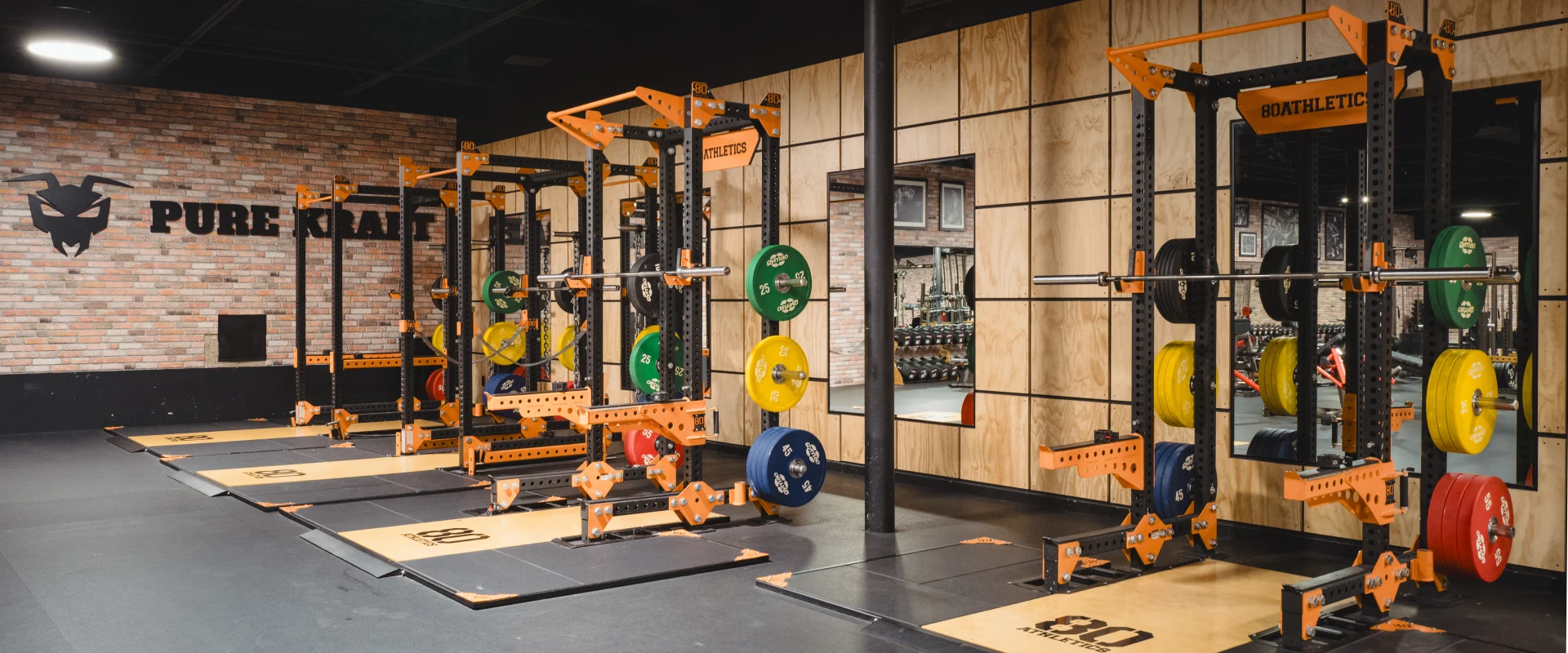 Power racks at Gold’s Gym Highland Meadows featuring orange frames and weight plates in multiple colors. The equipment sits on a rubber floor under ceiling lights, with a brick wall and the "Pure Kraft" logo in the background.