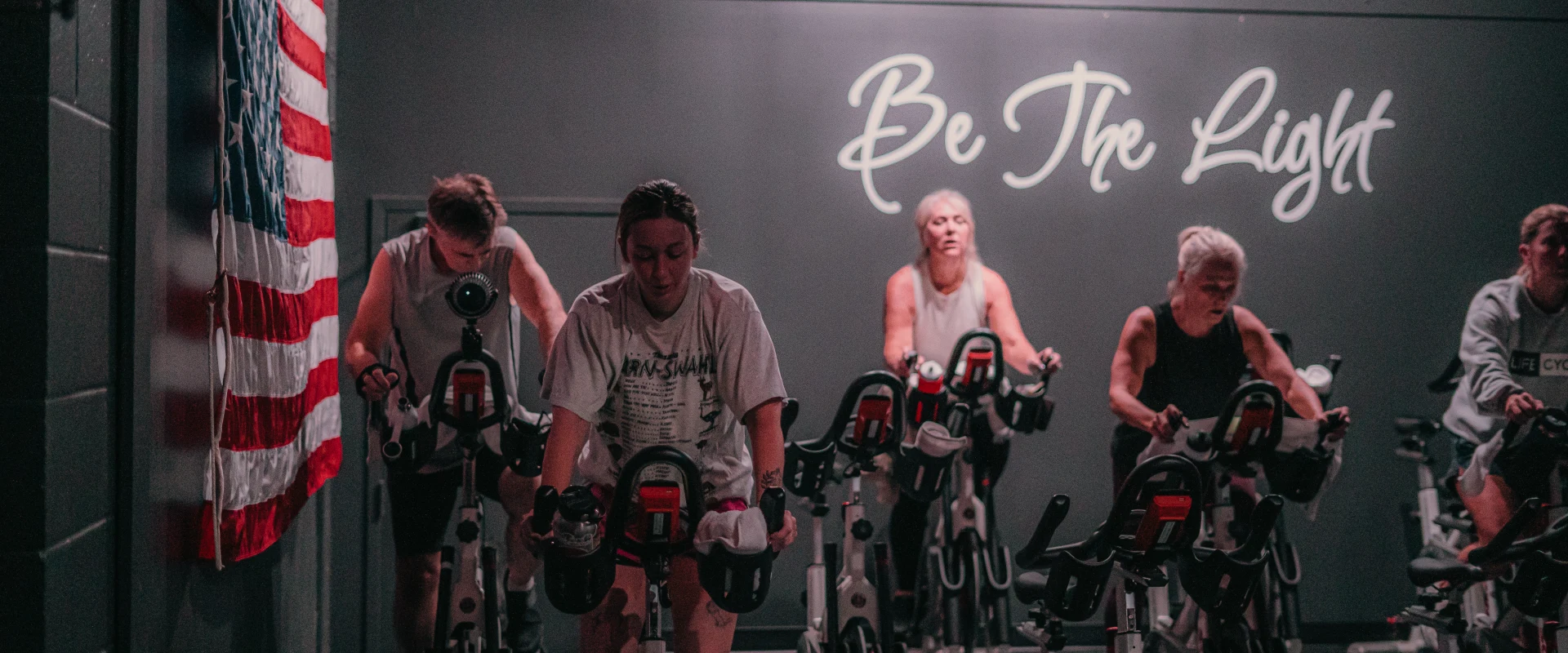 Group cycling class at Gold's Gym Hendersonville with participants riding stationary bikes in a dimly lit room featuring an American flag and the phrase "Be The Light" on the back wall.