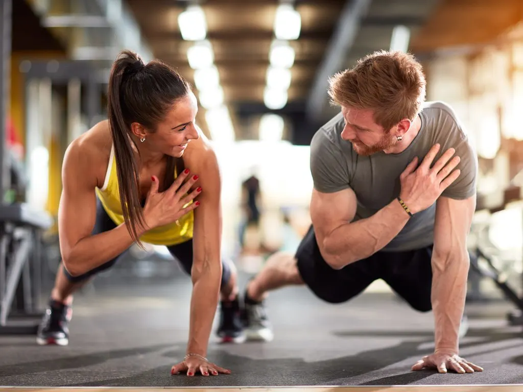 Two people, a woman and a man, are engaged in a fitness activity together in what appears to be a gym or exercise facility. The woman is in a yoga pose while the man is supporting her, creating an intimate and collaborative scene.