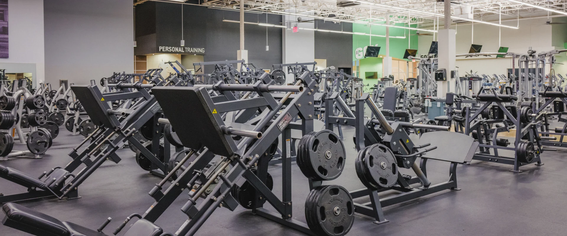 Free weights area at Gold's Gym Waxahachie featuring plate-loaded leg press machines and a variety of weightlifting equipment. The space has rubber flooring, bright overhead lighting creating a functional and spacious workout environment.