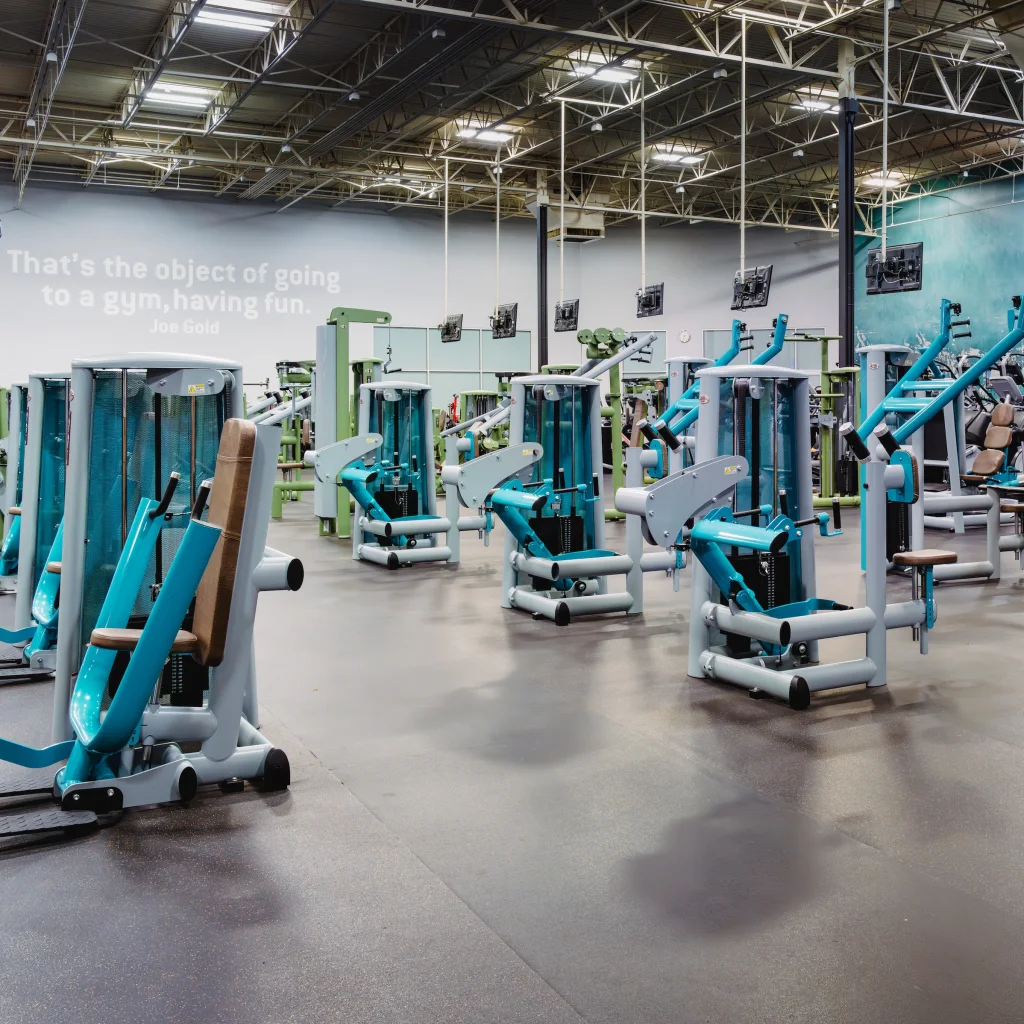 Wide gym floor with multiple strength machines under high ceilings and bright lighting.