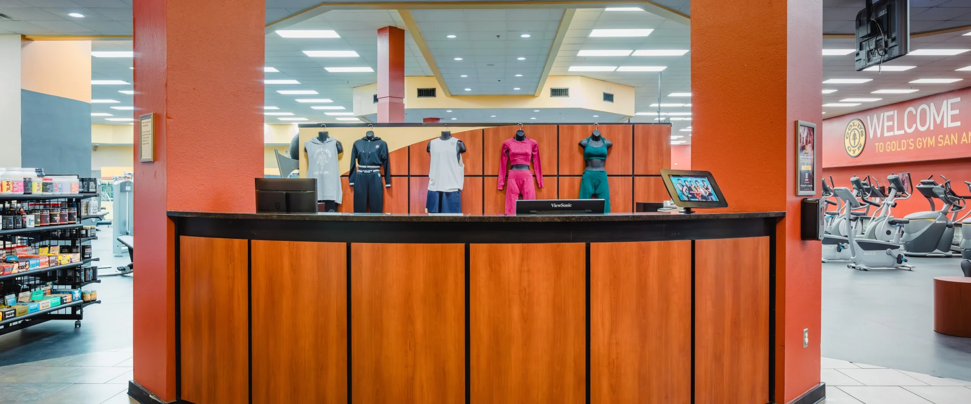 A gym entrance features a wooden reception desk, mannequins in sportswear, and a welcoming sign. Fitness equipment and shelves are visible, creating an energetic atmosphere.