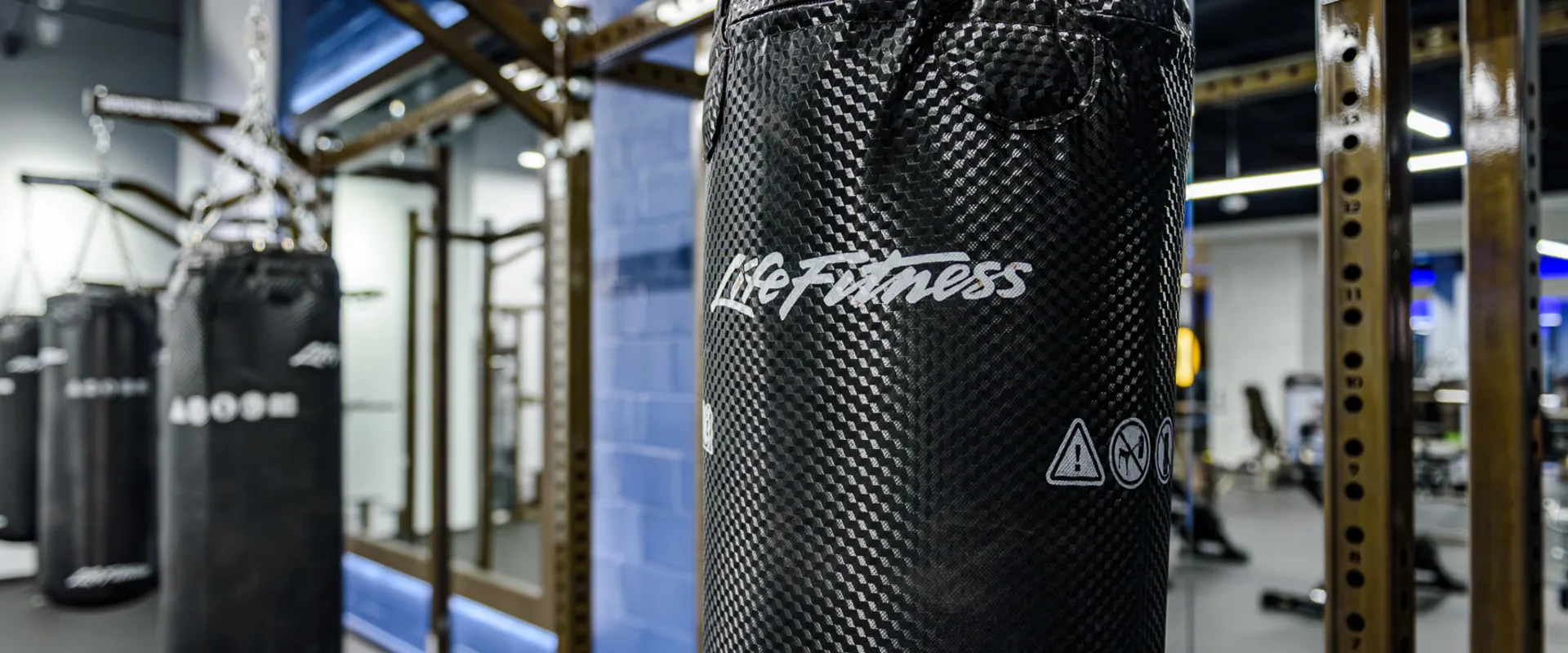 Boxing area at Gold’s Gym Thousand Oaks, featuring a close-up of a black Life Fitness punching bag with a textured surface, suspended by metal chains from a Hammer Strength frame. Additional punching bags hang in the background, reflected in large mirrors mounted on blue accent walls. Overhead LED lighting illuminates the space, with gym equipment visible beyond the boxing area. The floor is covered in rubber gym flooring for durability and support.