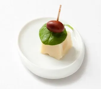 A white plate holds a small cube of cheese topped with a red cherry and a green leaf, creating a simple yet visually appealing presentation.