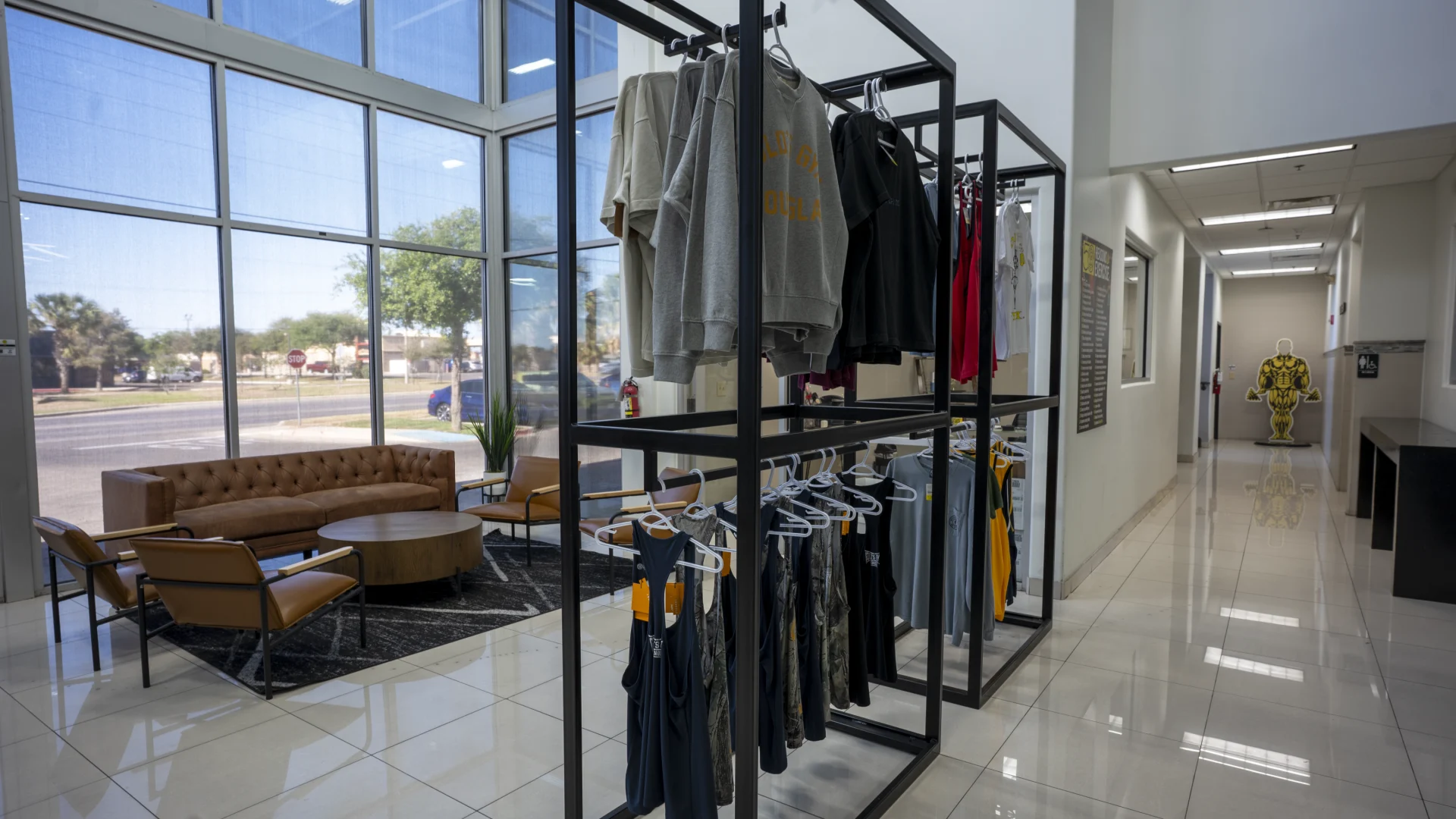 An interior view of a gym lobby featuring tall industrial black metal retail racks holding various apparel items like sweatshirts and tank tops. To the left is a lounge area with a brown leather tufted sofa, two matching armchairs, and a round wooden coffee table on a geometric rug, all set against floor-to-ceiling windows. A brightly lit hallway on the right leads toward a back area with a yellow bodybuilding statue visible in the distance.