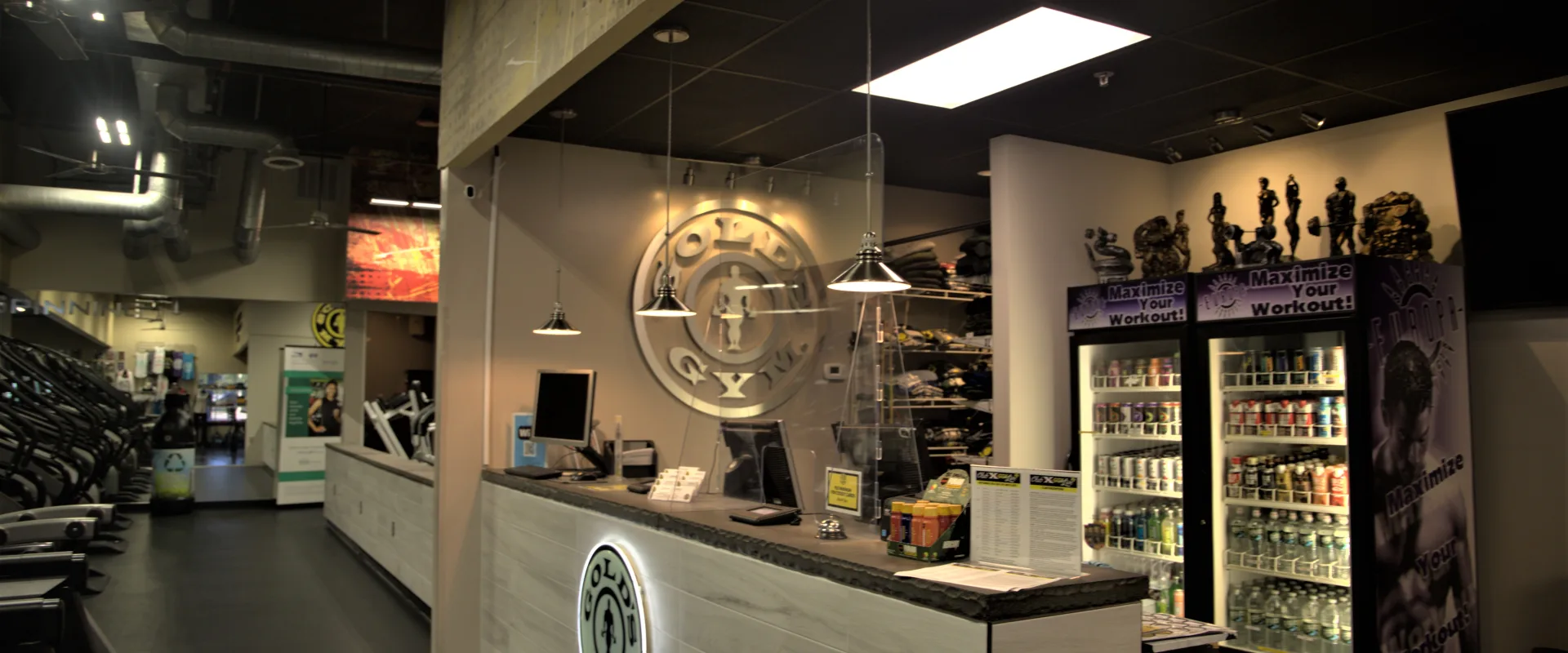 Gold's Gym Bridgewater front desk area featuring a modern reception counter with the gym's logo illuminated on the front panel. Pendant lights hang from the ceiling, highlighting the counter with computers. A refrigerated display stocked with fitness drinks and supplements stands to the side, along with gym apparel and trophies on shelves in the background. The open space leads into the cardio section with treadmills visible in the distance.