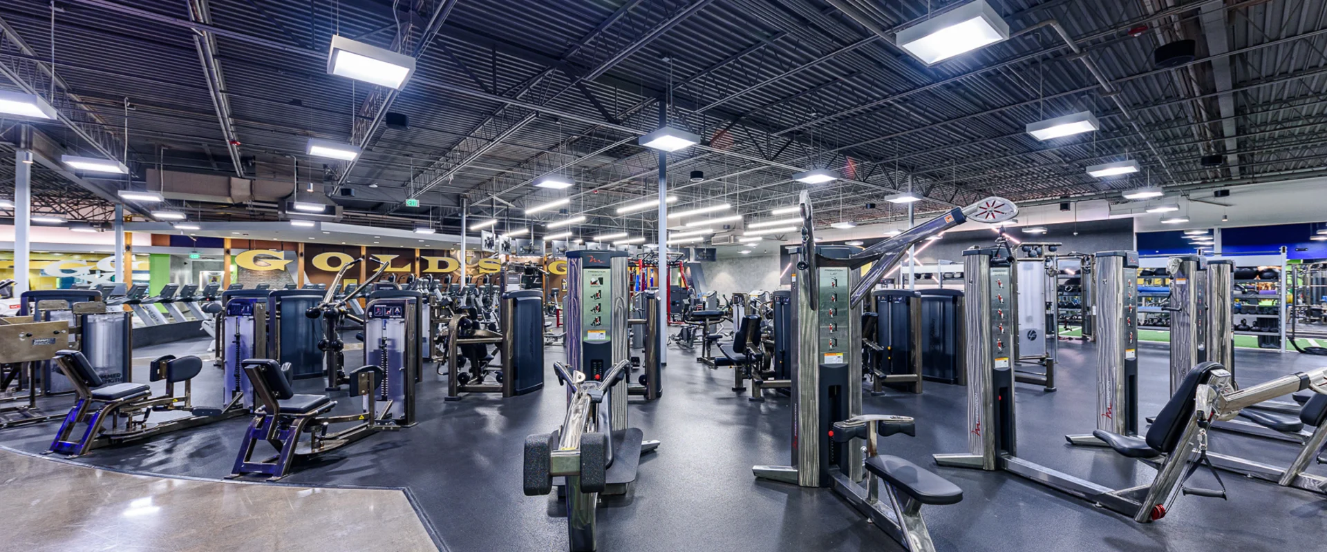 Strength training area at Gold's Gym Garden Grove, showcasing a variety of weight machines arranged in a spacious, open floor plan. Machines are polished chrome and navy blue, set on a mix of rubber and polished concrete flooring. Overhead lighting and mirrors enhance visibility, while the Gold’s Gym logo decorates the far wall.