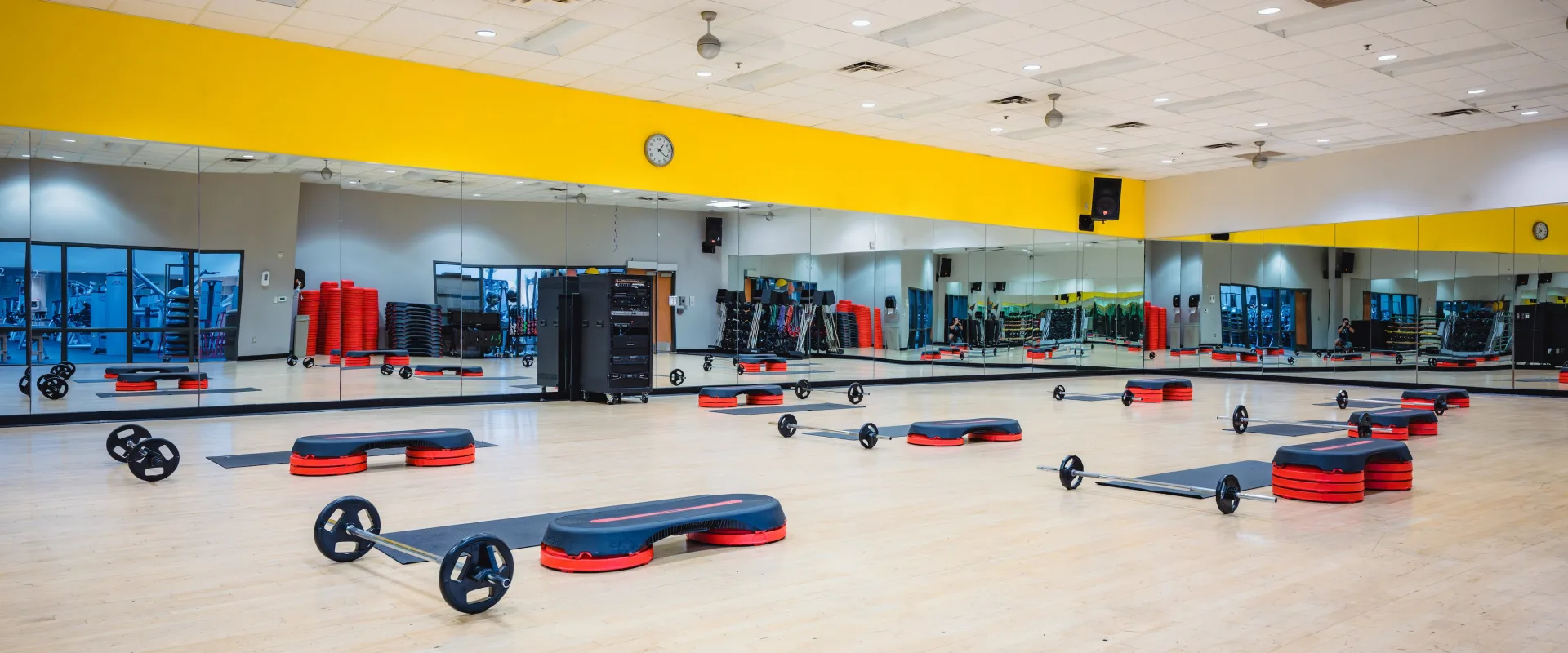 Brightly lit group exercise room with mirrored walls and a yellow accent. Exercise steps, weights, and bars are neatly arranged on the wooden floor.