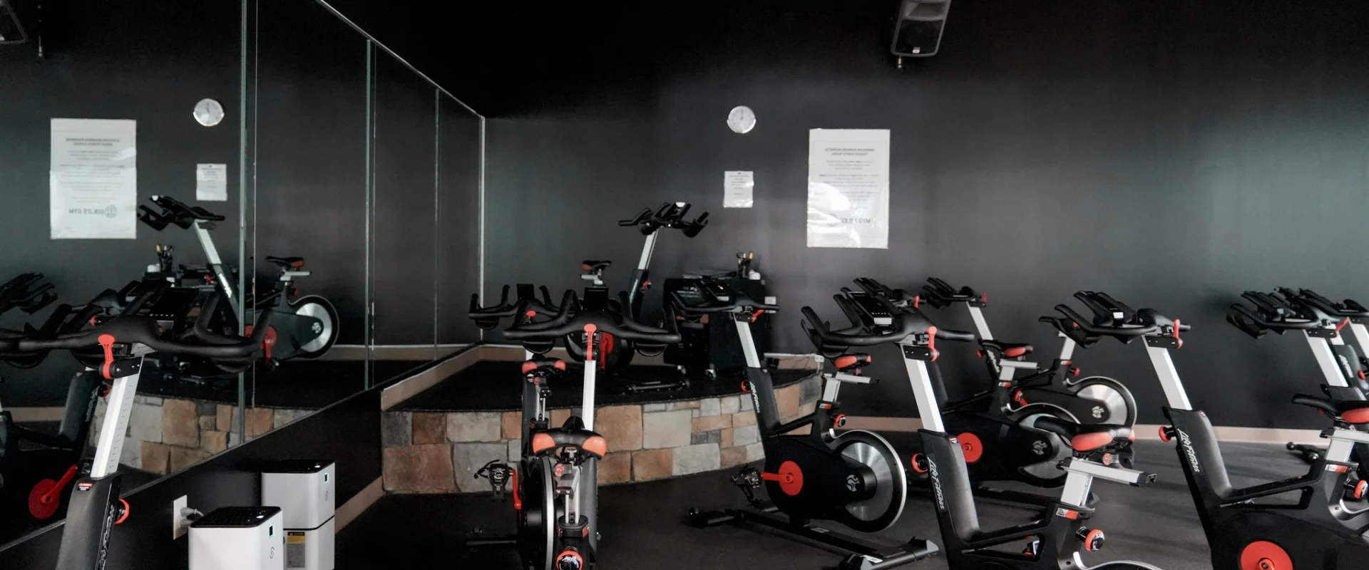 A dimly lit indoor cycling room at Gold's Gym Rock Hill featuring multiple stationary bikes arranged in rows. A raised instructor’s platform sits against a mirrored wall, with black walls and subtle lighting creating a focused workout environment. Large ceiling fans and wall-mounted speakers enhance the class experience.