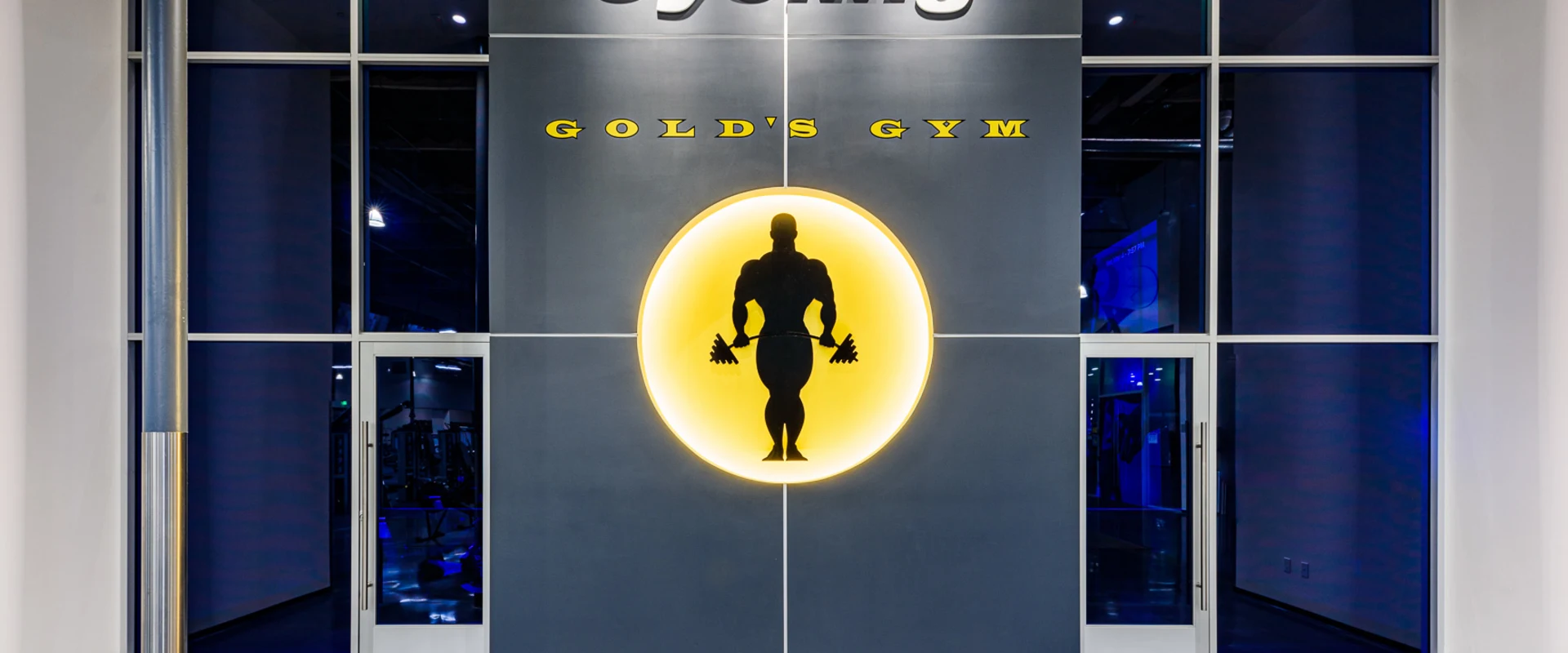 The group cycle studio at Gold’s Gym Cerritos features a dark gray accent wall with the word 'Cycling' in bold white letters and the Gold’s Gym logo illuminated in yellow. Floor-to-ceiling windows with glass doors provide a view into the studio, reflecting blue ambient lighting from inside. The polished concrete floor and a stainless steel support column add to the modern aesthetic.