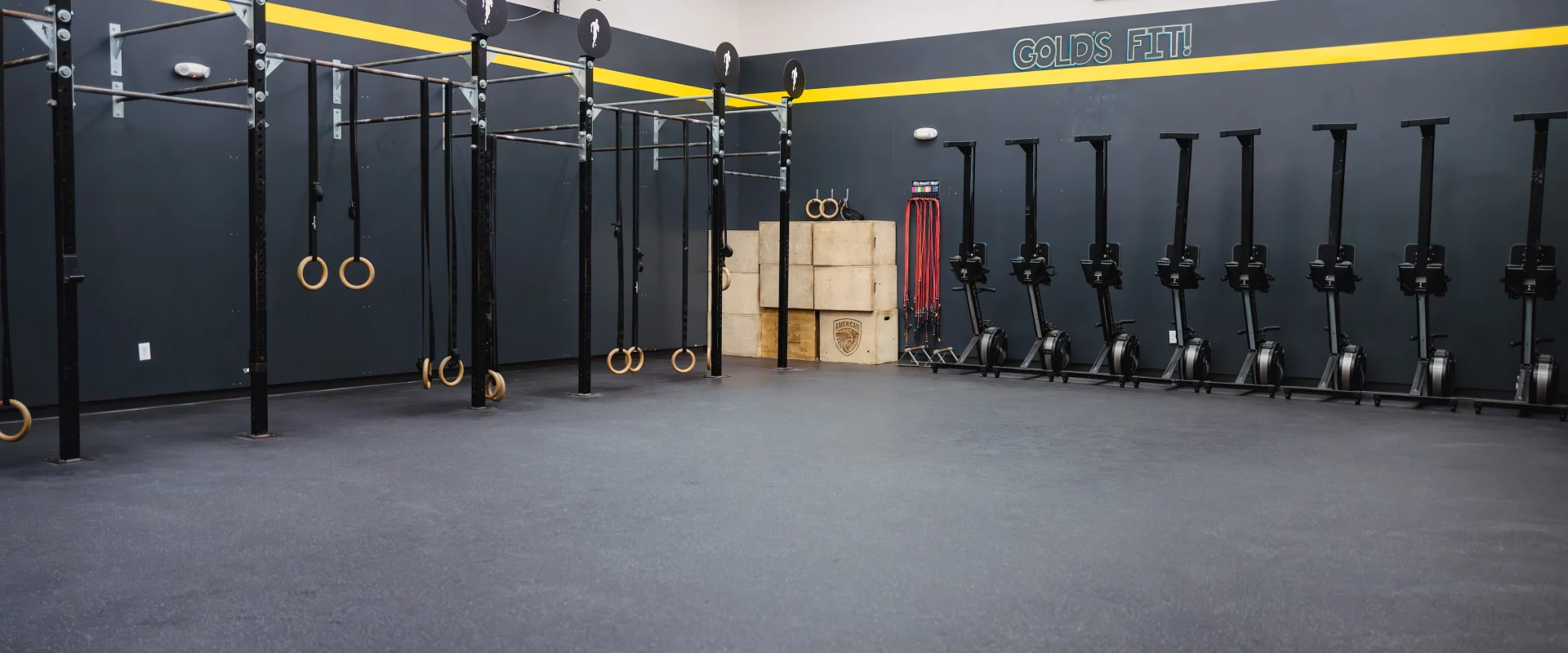 Spacious gym with black-and-yellow walls, pull-up bars, gymnastic rings, stacked boxes, and row machines lined up, creating a focused workout vibe.