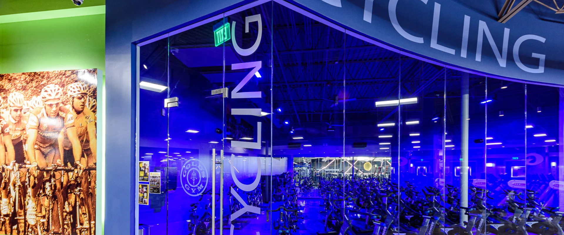 The dedicated group cycling studio is enclosed by curved floor-to-ceiling glass panels with "Cycling" lettering, illuminated in blue lighting. A large image of cyclists in a race decorates the adjacent green wall.