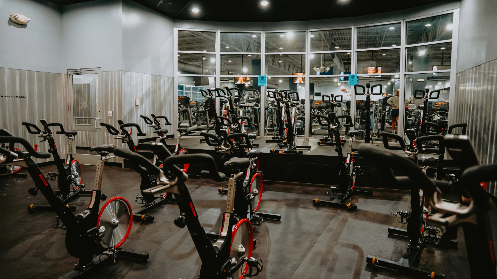 Group cycle studio at Gold’s Gym Tennessee, Lee Highway, featuring rows of stationary bikes arranged on a dark rubber floor. The space includes a raised platform for the instructor and large windows in the background providing a view of the gym floor. The room is illuminated by overhead ceiling lighting, creating a bright environment for cycling classes.