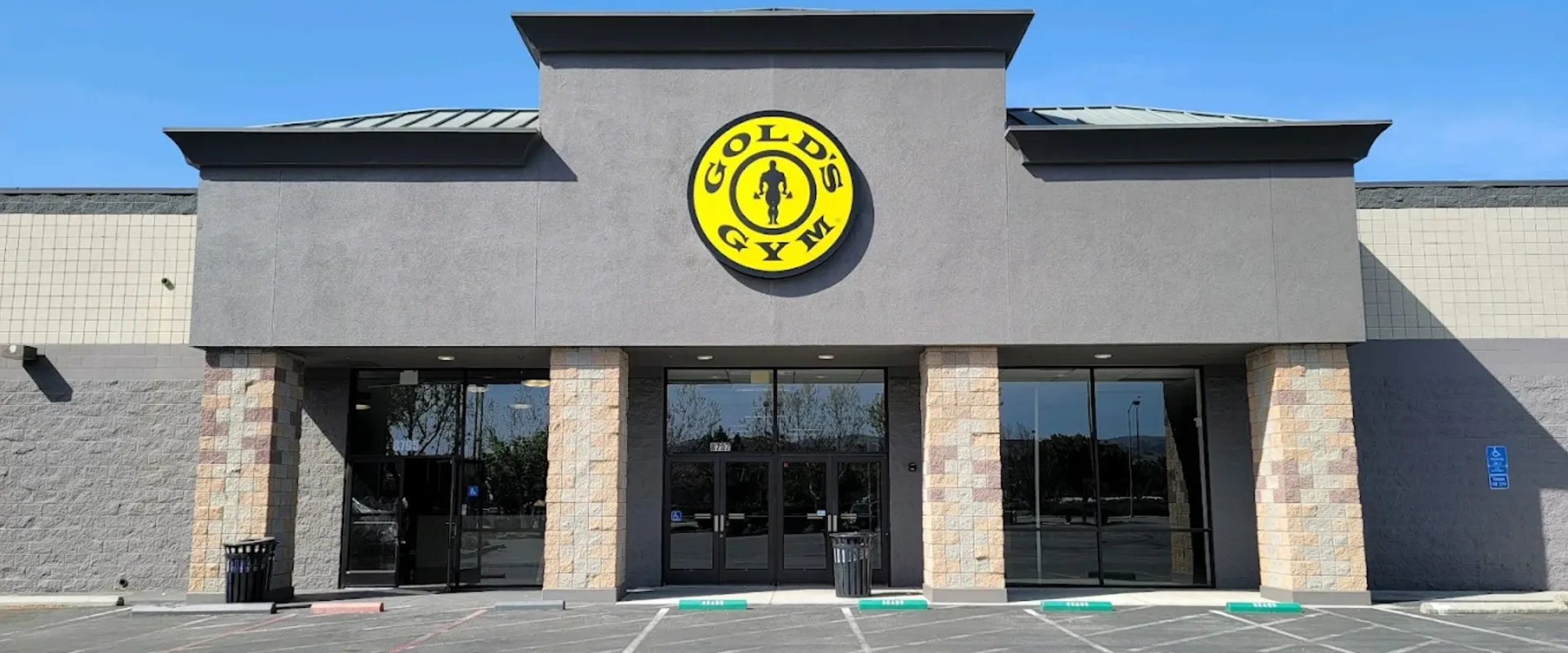 Exterior of Gold’s Gym Gilroy with yellow circular logo above entrance on gray and tan building under blue sky.
