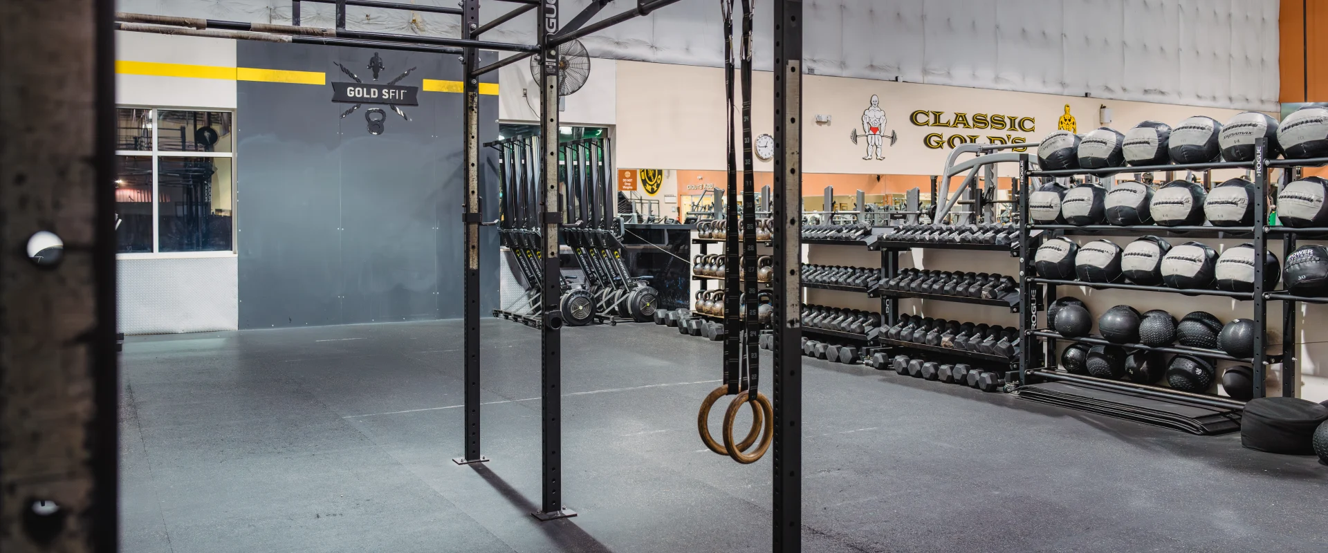 Gold's Fit area at Gold's Gym Austin (Bee Caves) featuring a functional training rig with gymnastic rings and pull-up bars. Surrounding the rig are racks of medicine balls, kettlebells, and other functional fitness equipment. The space has black rubber flooring, with "Gold's Fit" branding on the wall.