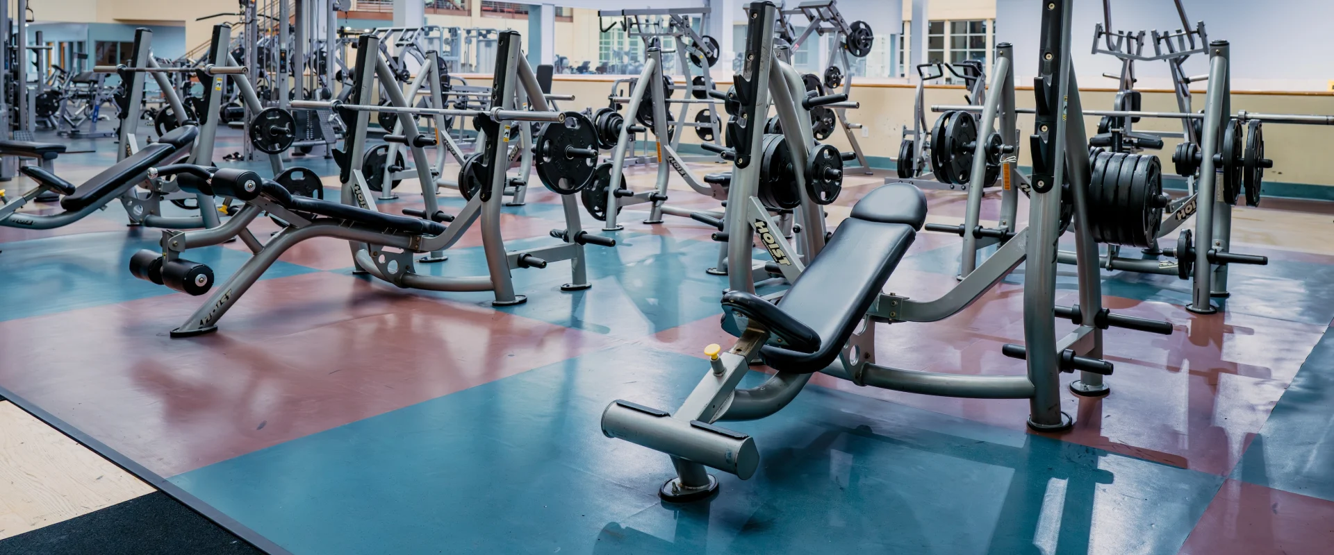 Spacious gym with various weightlifting equipment, including benches, barbells, and weight racks. The environment is well-lit and orderly.