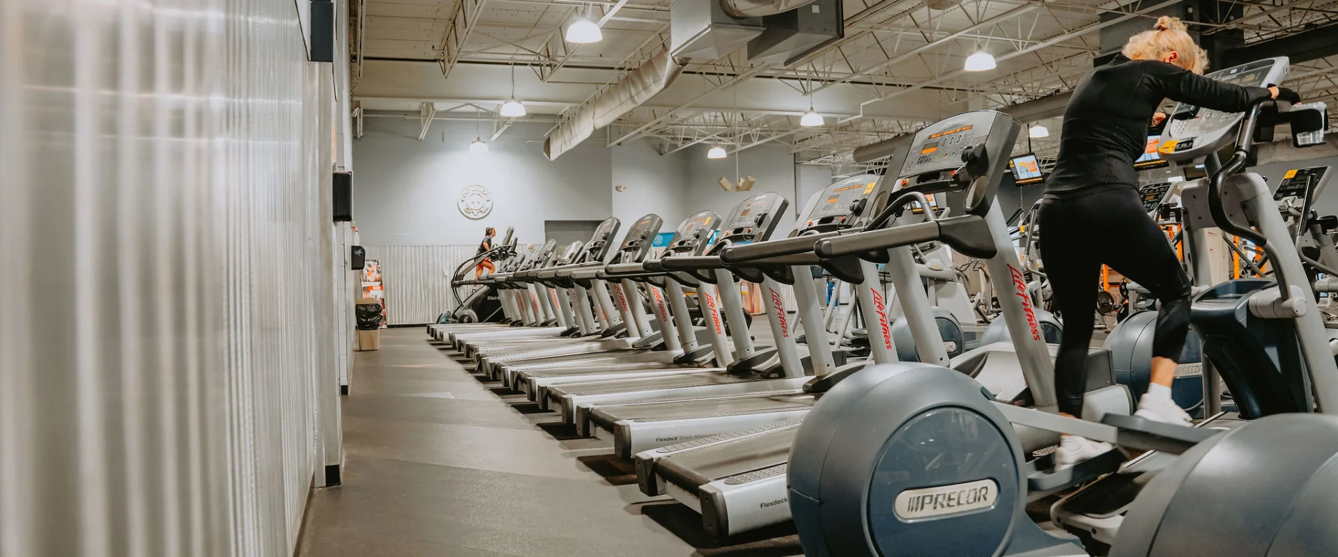 Cardio equipment area at Gold’s Gym Tennessee, Hixson, featuring a row of treadmills and elliptical machines on a dark rubberized floor. The space is illuminated by high ceilings with exposed white ducts and industrial-style overhead lighting. A member is seen using an elliptical in the foreground, with a sleek corrugated metal wall running along the left side. The gym's signature logo is displayed on a gray accent wall in the background, adding a touch of branding to the spacious and clean environment.